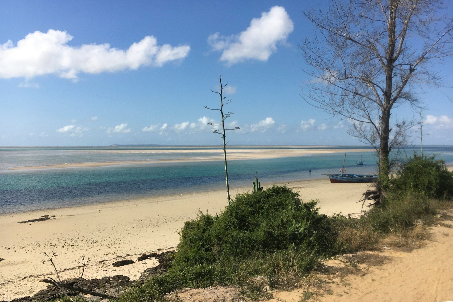 Una barca riposa su una spiaggia sabbiosa accanto ad acque turchesi poco profonde e banchi di sabbia sotto un cielo azzurro con nuvole sparse.