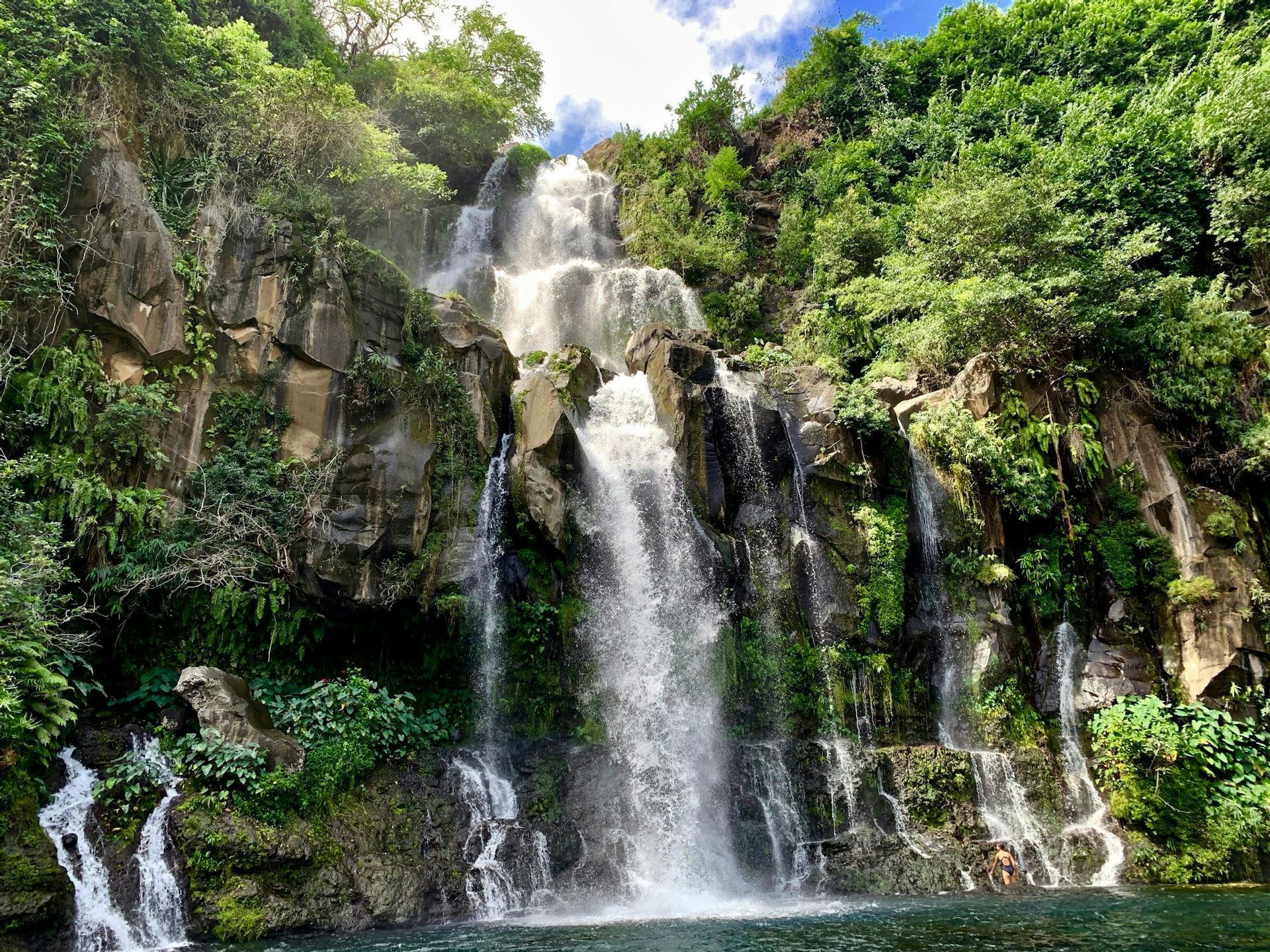 Una cascata a più livelli si getta da una lussureggiante parete rocciosa in una piscina naturale sottostante, con una persona seduta nell'acqua.