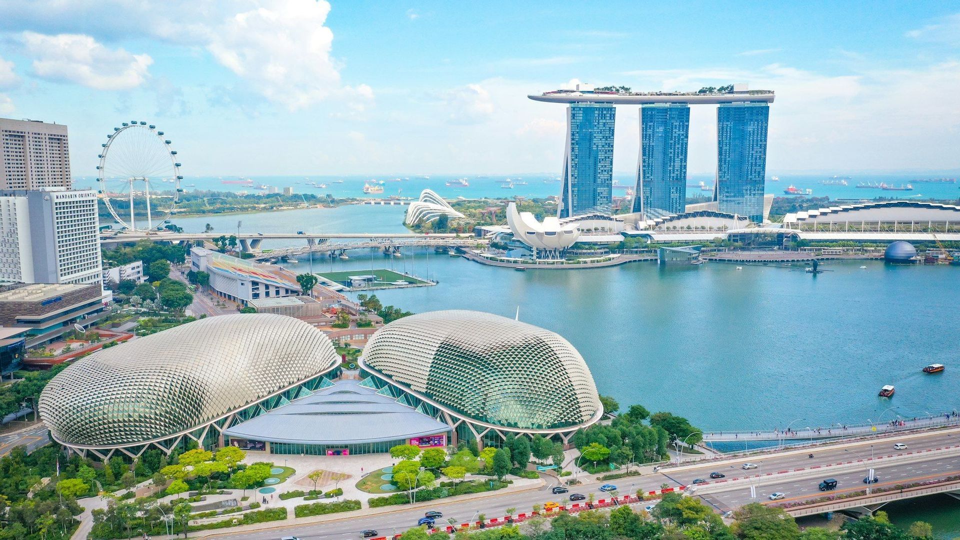 Una veduta aerea del lungomare di una città moderna, con edifici dall'architettura unica, una grande ruota panoramica e una baia trafficata sotto un cielo parzialmente nuvoloso.