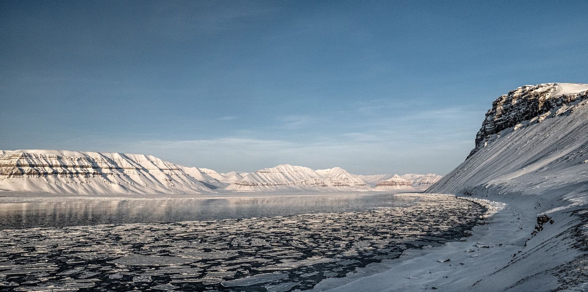 Un fiordo artico pieno di lastre di ghiaccio galleggianti è circondato da montagne innevate sotto un cielo blu chiaro.