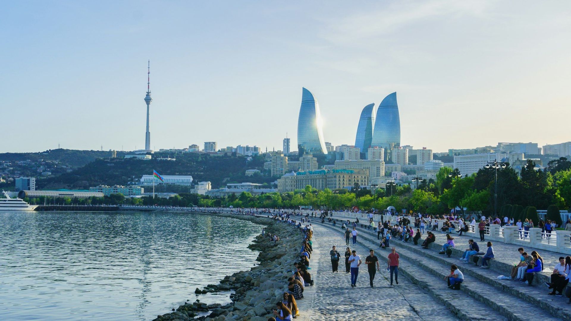 Menschen spazieren und entspannen sich auf einer breiten Steinpromenade am Wasser, vor einer modernen Stadt-Skyline mit markanten Glastürmen.