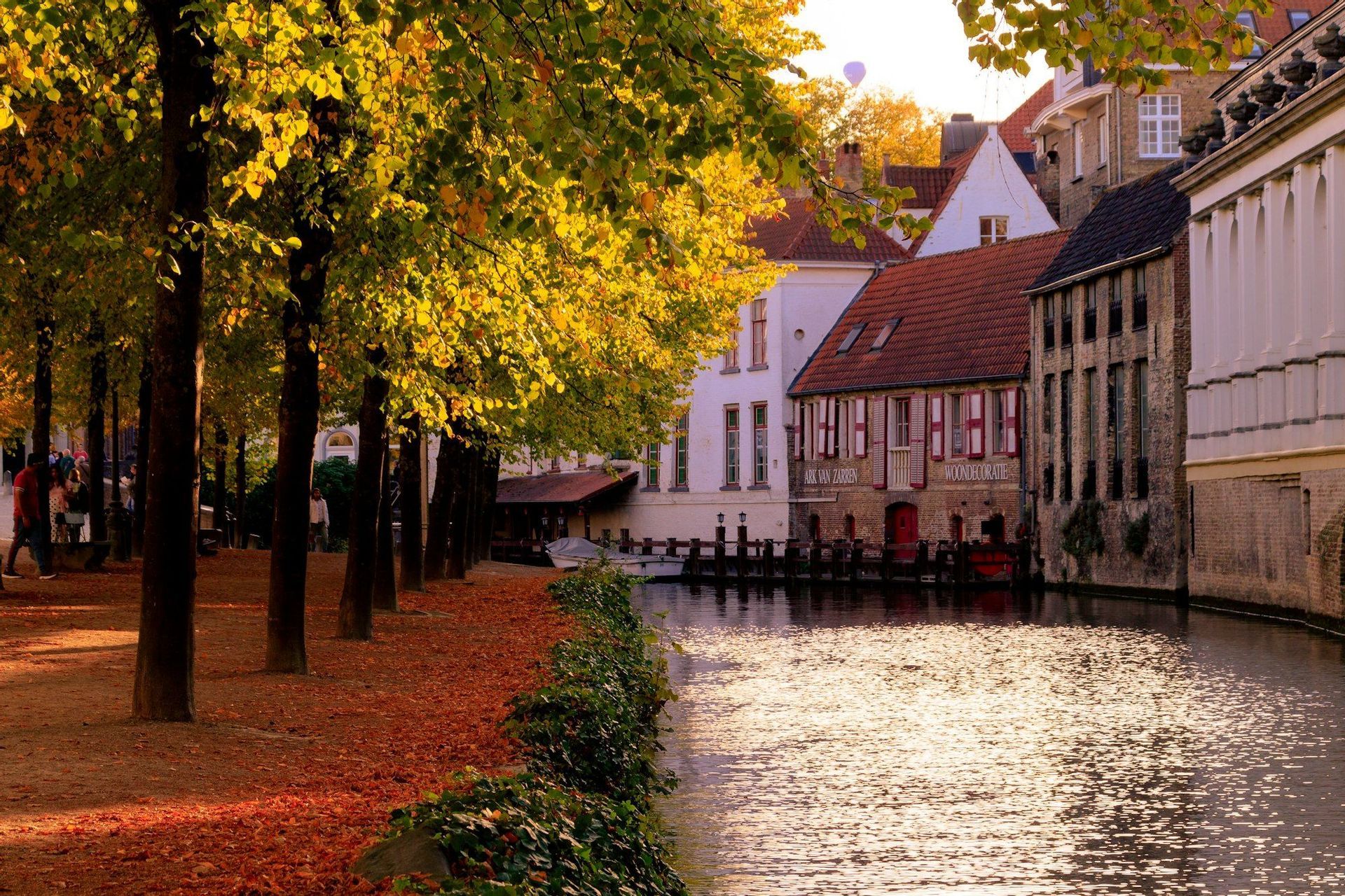 Ein sonnenbeschienener Kanal im Herbst, auf einer Seite von historischen Gebäuden und auf der anderen von einem baumgesäumten Weg mit gefallenen roten Blättern gesäumt.