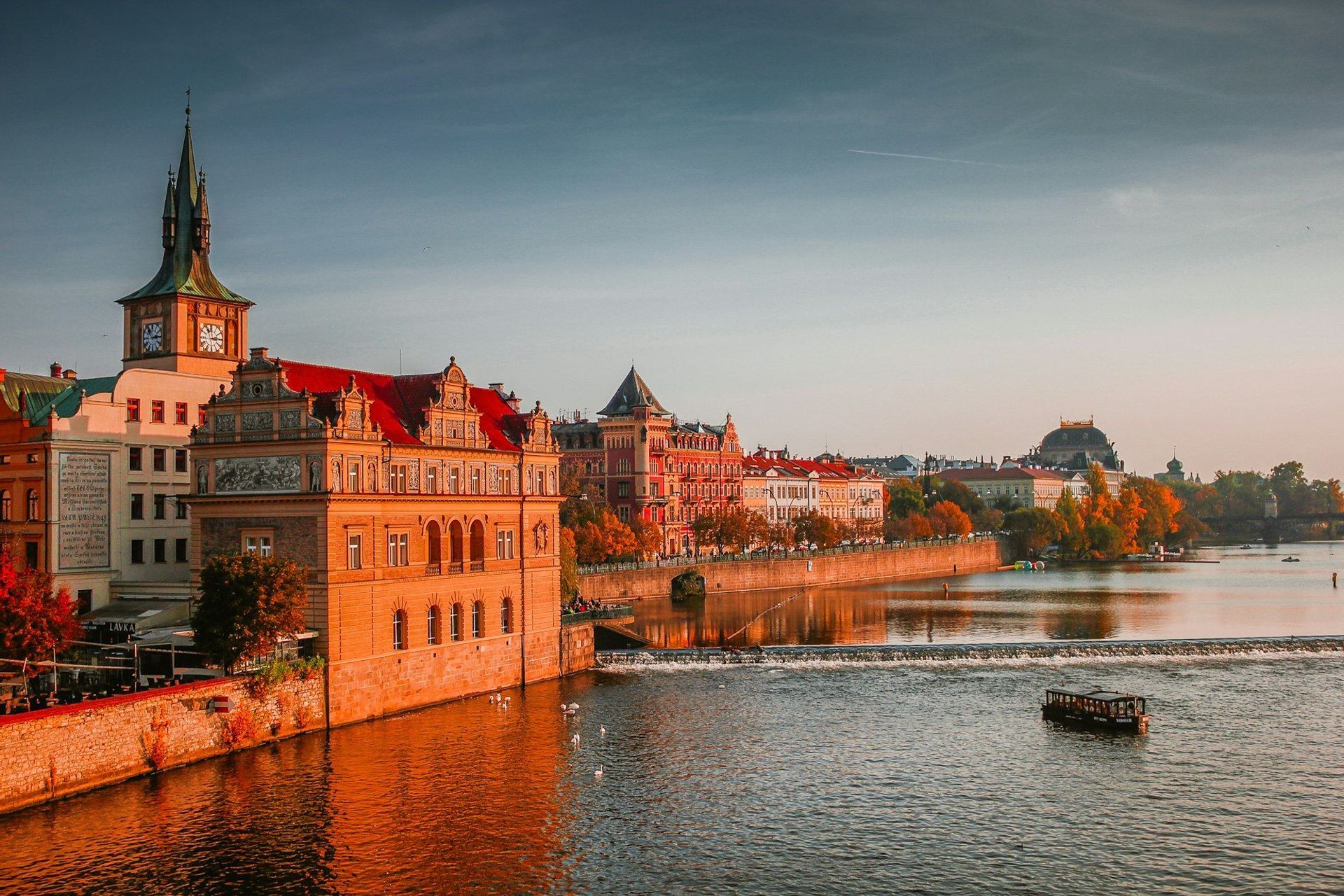 Historische Gebäude mit roten Dächern und einem Uhrturm säumen einen breiten Fluss während eines goldenen Sonnenuntergangs im Herbst.