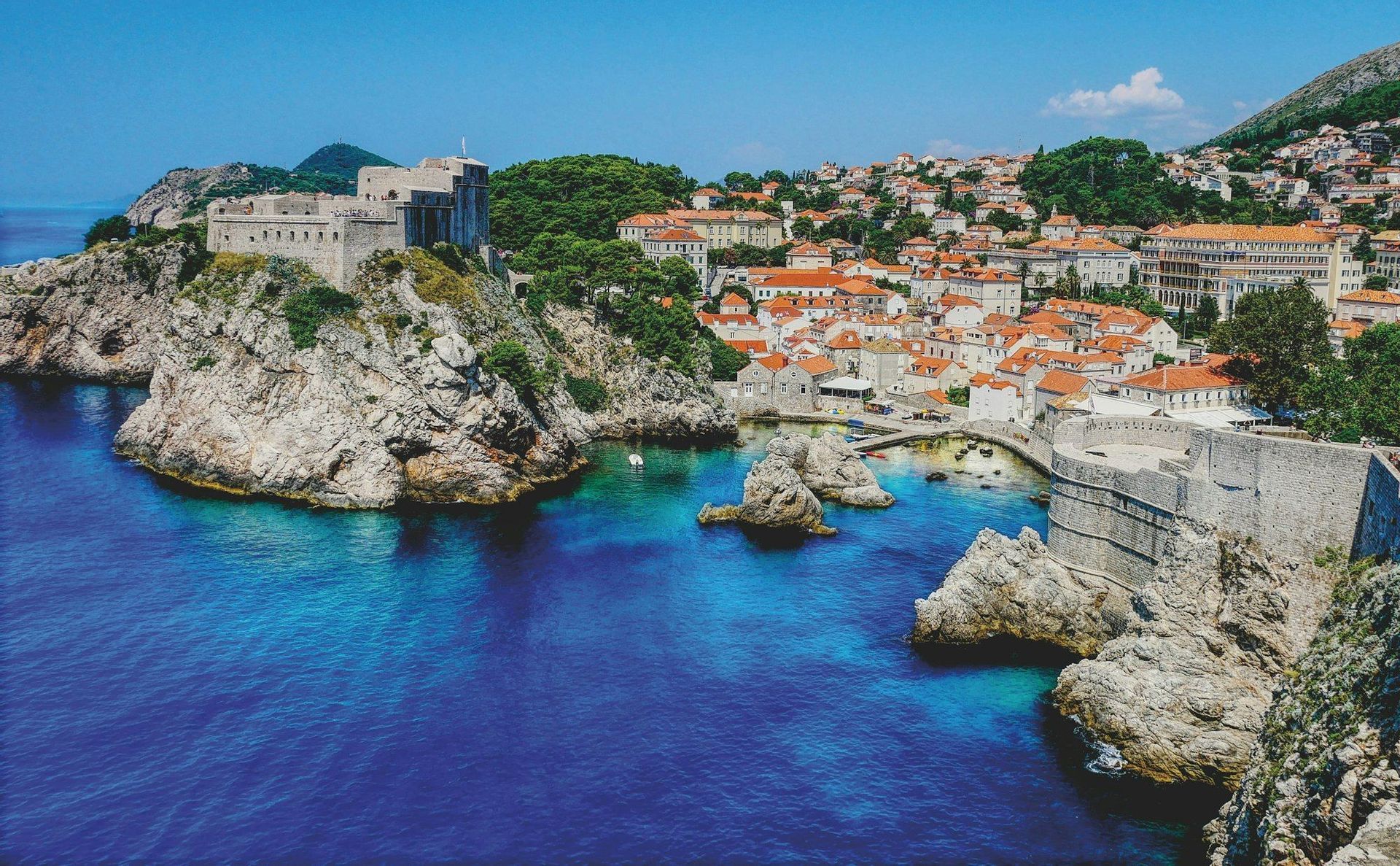 Una fortezza storica e una città fortificata con tetti in terracotta si trovano su una costa rocciosa accanto a un mare blu brillante.