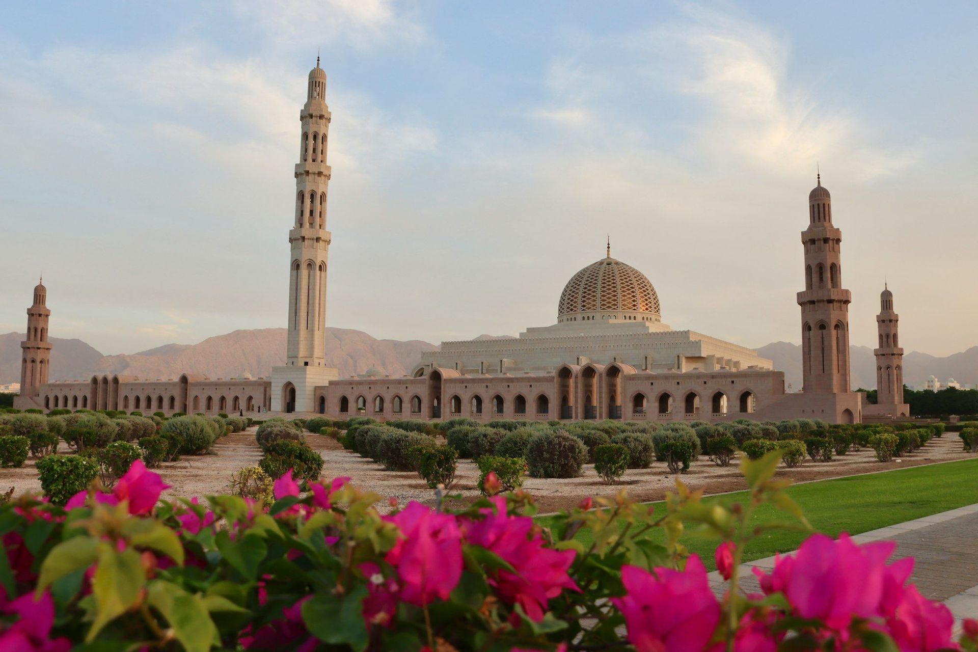 Hinter einem gepflegten Garten mit rosa Blumen im Vordergrund und Bergen im Hintergrund steht eine große Moschee mit zentraler Kuppel und Minaretten.