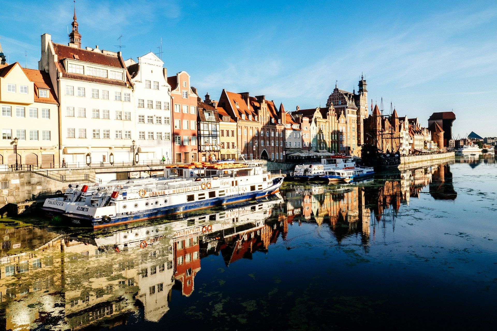Bunte historische Gebäude und am Fluss liegende Boote spiegeln sich im ruhigen Wasser unter klarem blauem Himmel.
