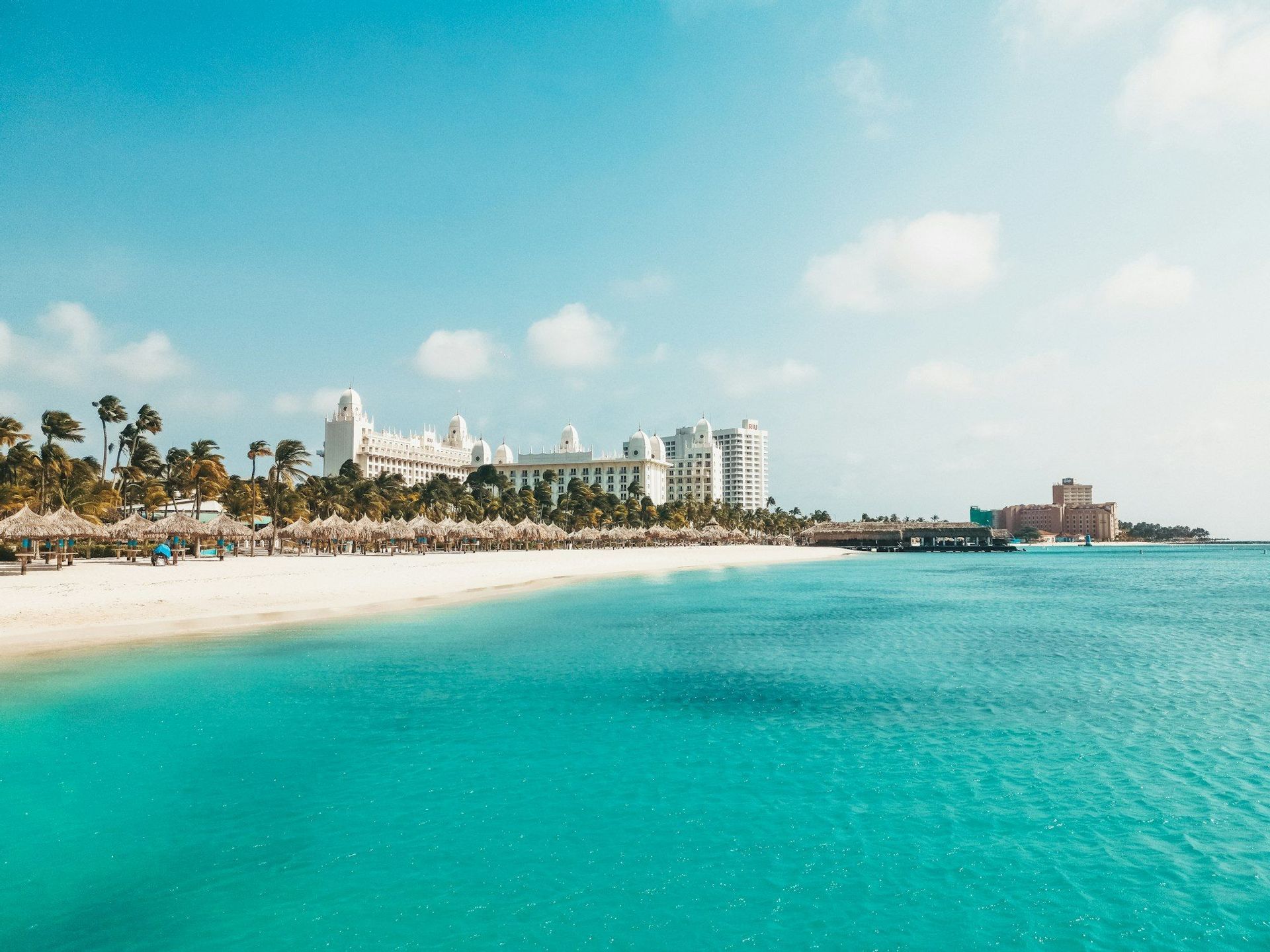 Una spiaggia ampia di sabbia bianca con palme e un grande hotel incontra l'oceano turchese sotto un cielo azzurro brillante.
