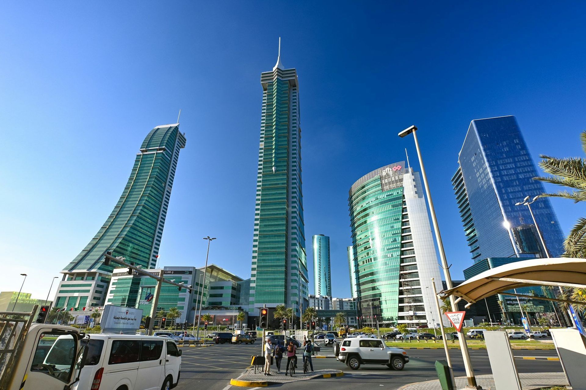 Alti grattacieli di vetro con architettura moderna dominano una trafficata strada cittadina, animata da auto e pedoni, sotto un cielo azzurro e limpido.
