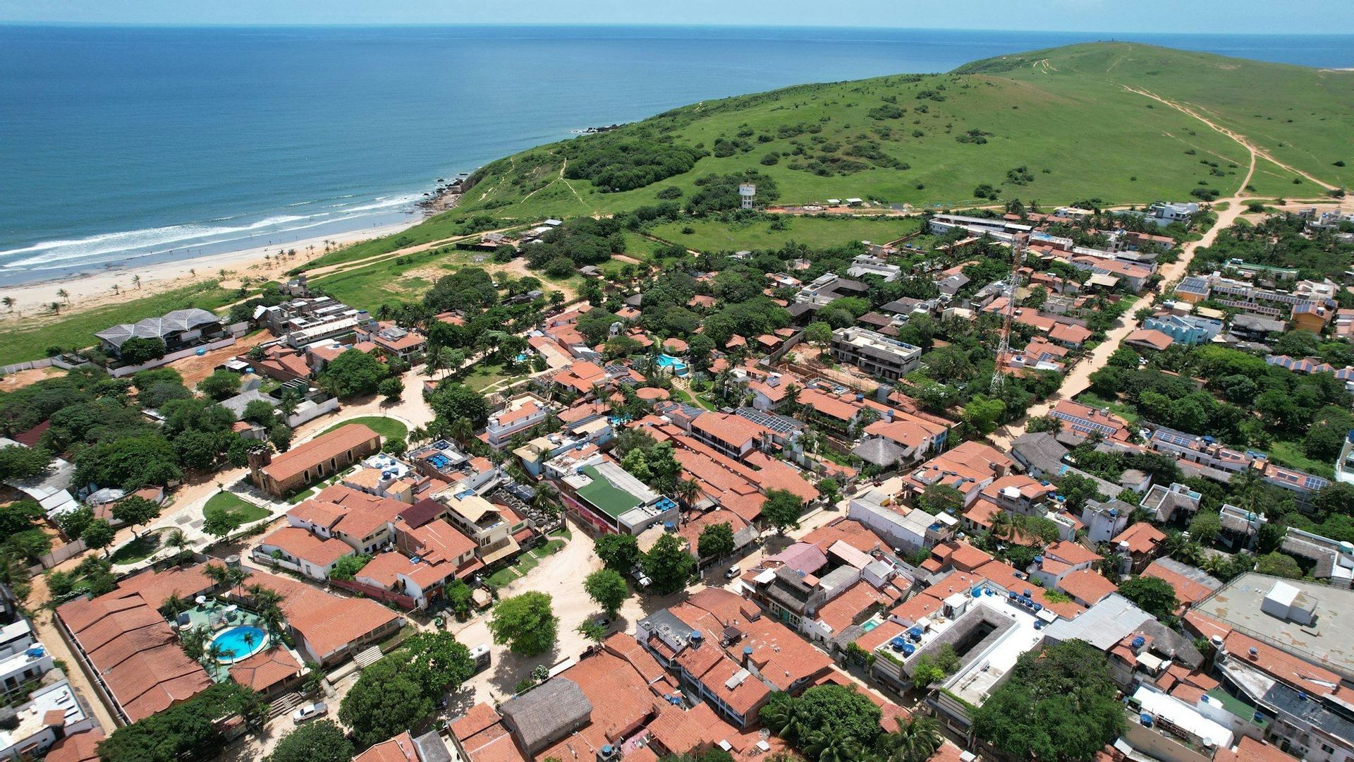 Una vista aerea di una città costiera con tetti in tegole rosse incastonata tra una collina erbosa e un oceano blu con una spiaggia sabbiosa.