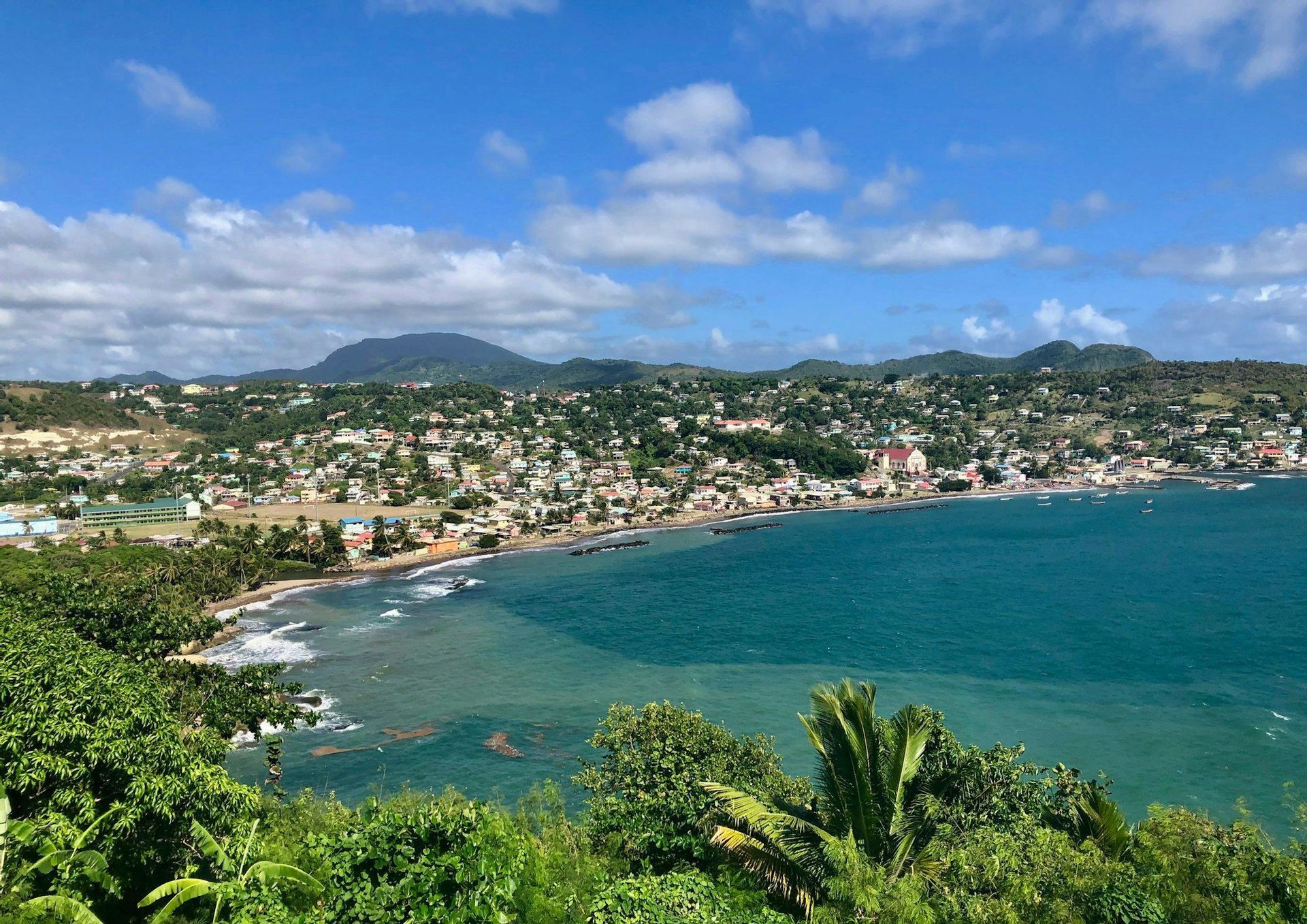 Una vista panoramica di una città costiera con edifici colorati adagiati in una baia, con verdi colline sullo sfondo sotto un cielo parzialmente nuvoloso.