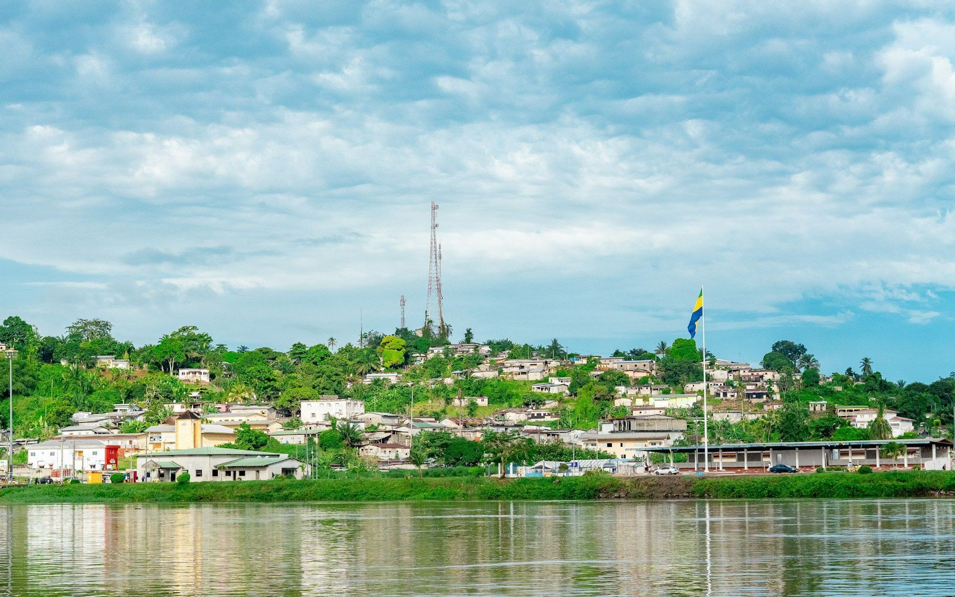Una vista da oltre un fiume di una città collinare coperta di alberi verdi lussureggianti, con la bandiera del Gabon che sventola sotto un cielo nuvoloso.