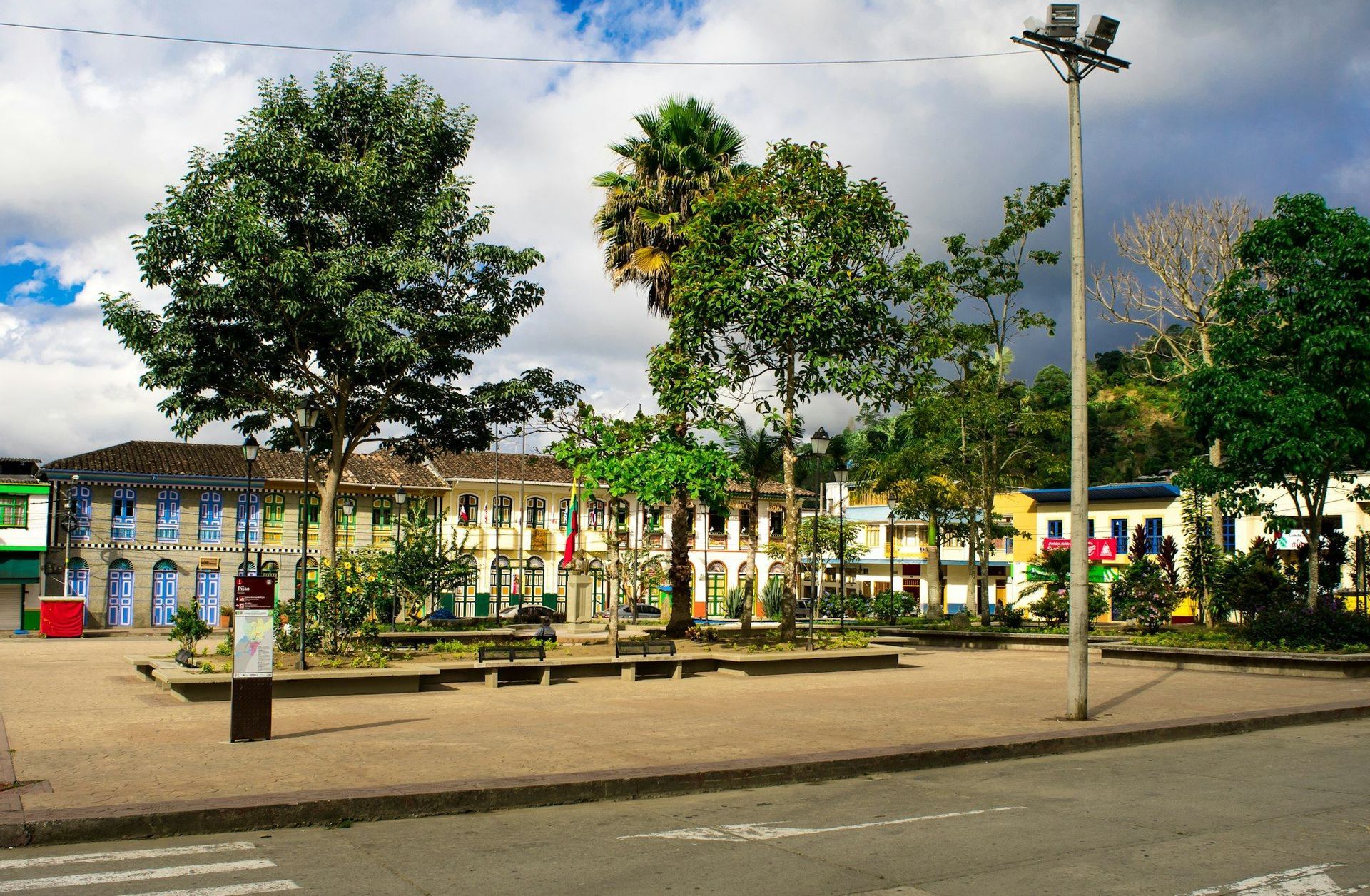 Una piazza cittadina pavimentata con alberi e panchine, circondata da colorati edifici in stile coloniale sotto un cielo parzialmente nuvoloso.
