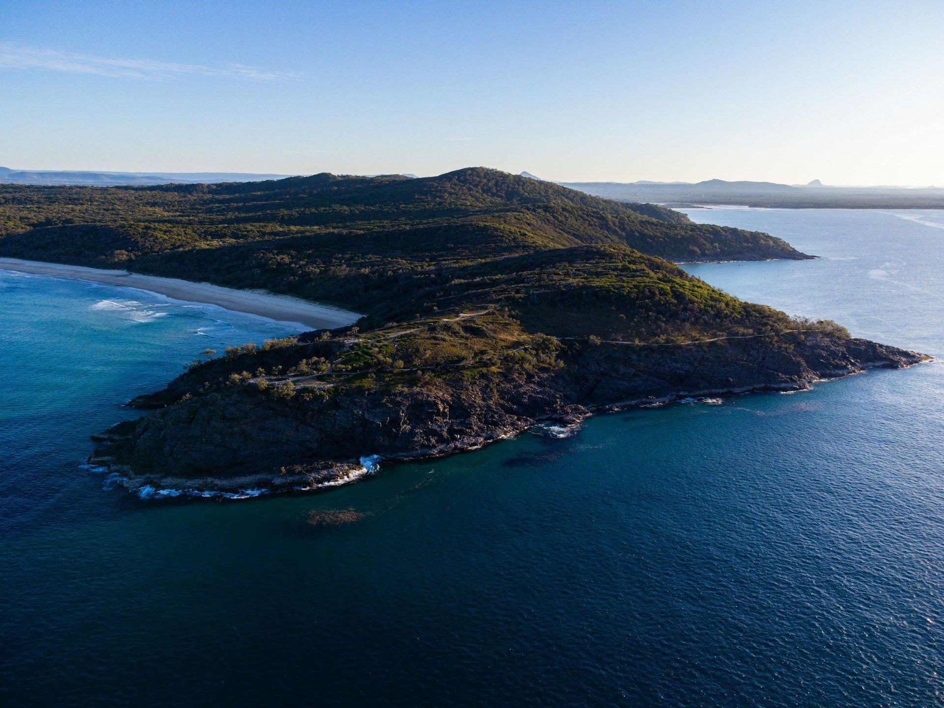 Una vista aerea di un promontorio verde e collinare che si estende nel profondo mare blu, con una spiaggia sabbiosa su un lato sotto un cielo sereno.