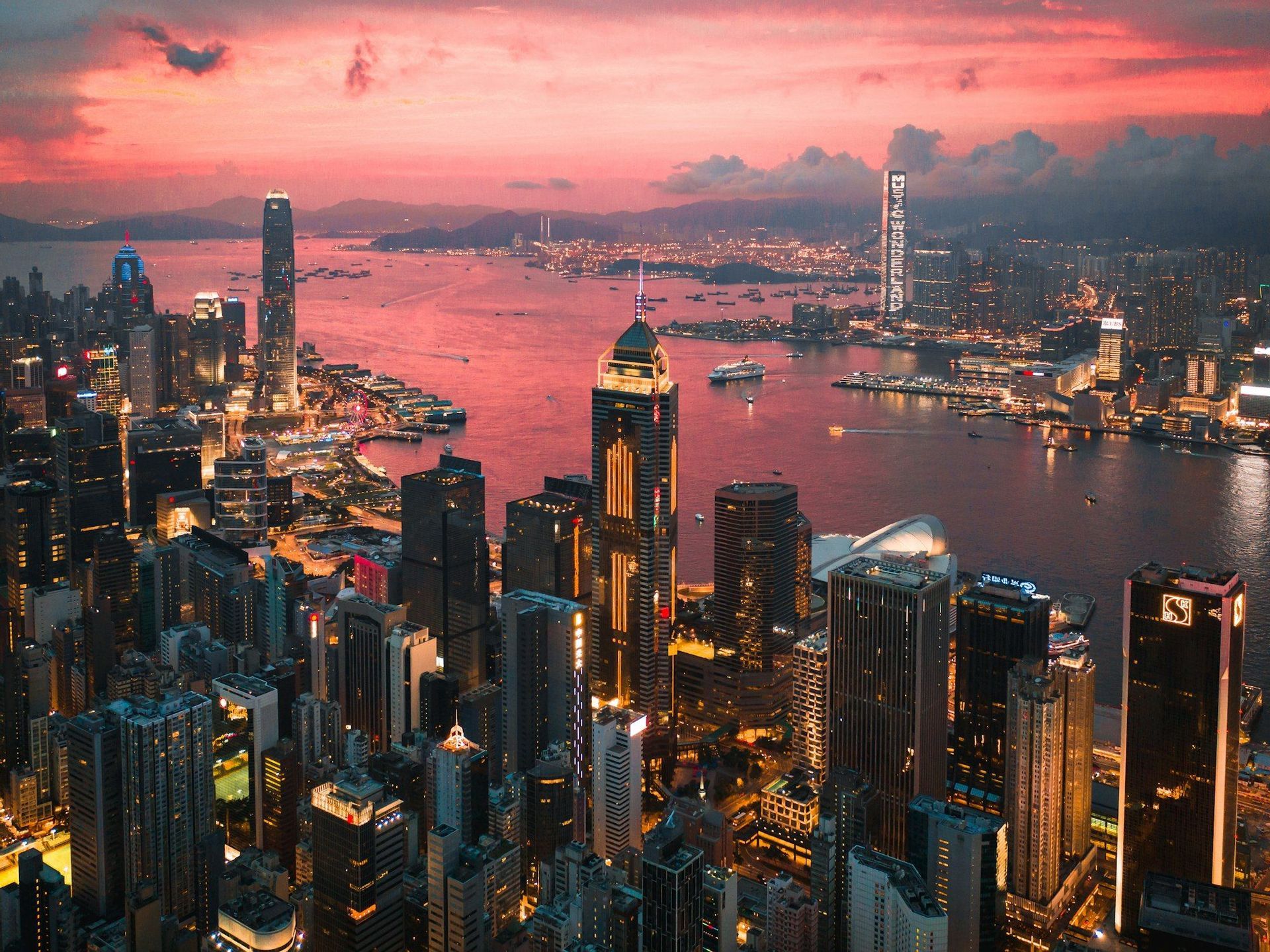 Veduta aerea di uno skyline cittadino illuminato con numerosi grattacieli lungo un lungomare, sotto un cielo rosa vibrante al tramonto.