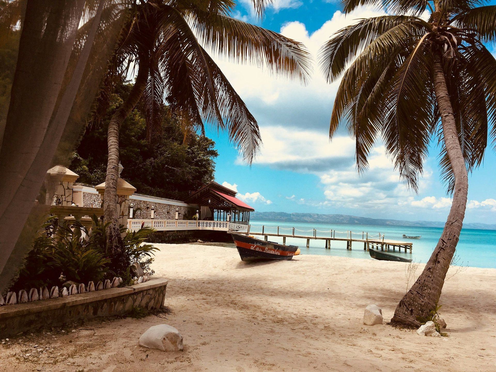 Palme su una spiaggia sabbiosa con piccole imbarcazioni e un pontile di legno che si estende in acque calme e turchesi sotto un cielo parzialmente nuvoloso.