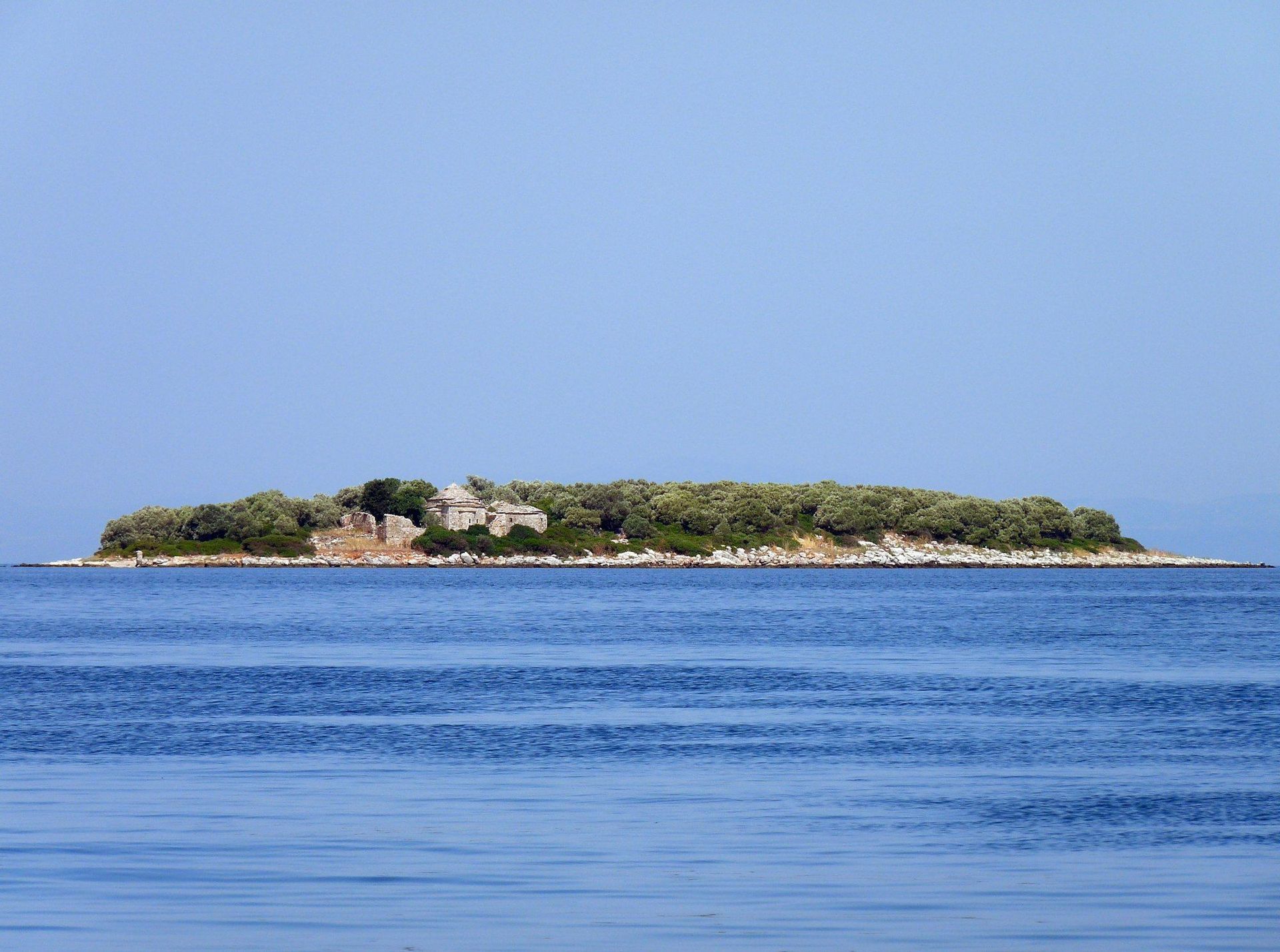 Una piccola isola allungata coperta di alberi verdi e antiche rovine in pietra, circondata da un mare blu calmo sotto un cielo sereno.