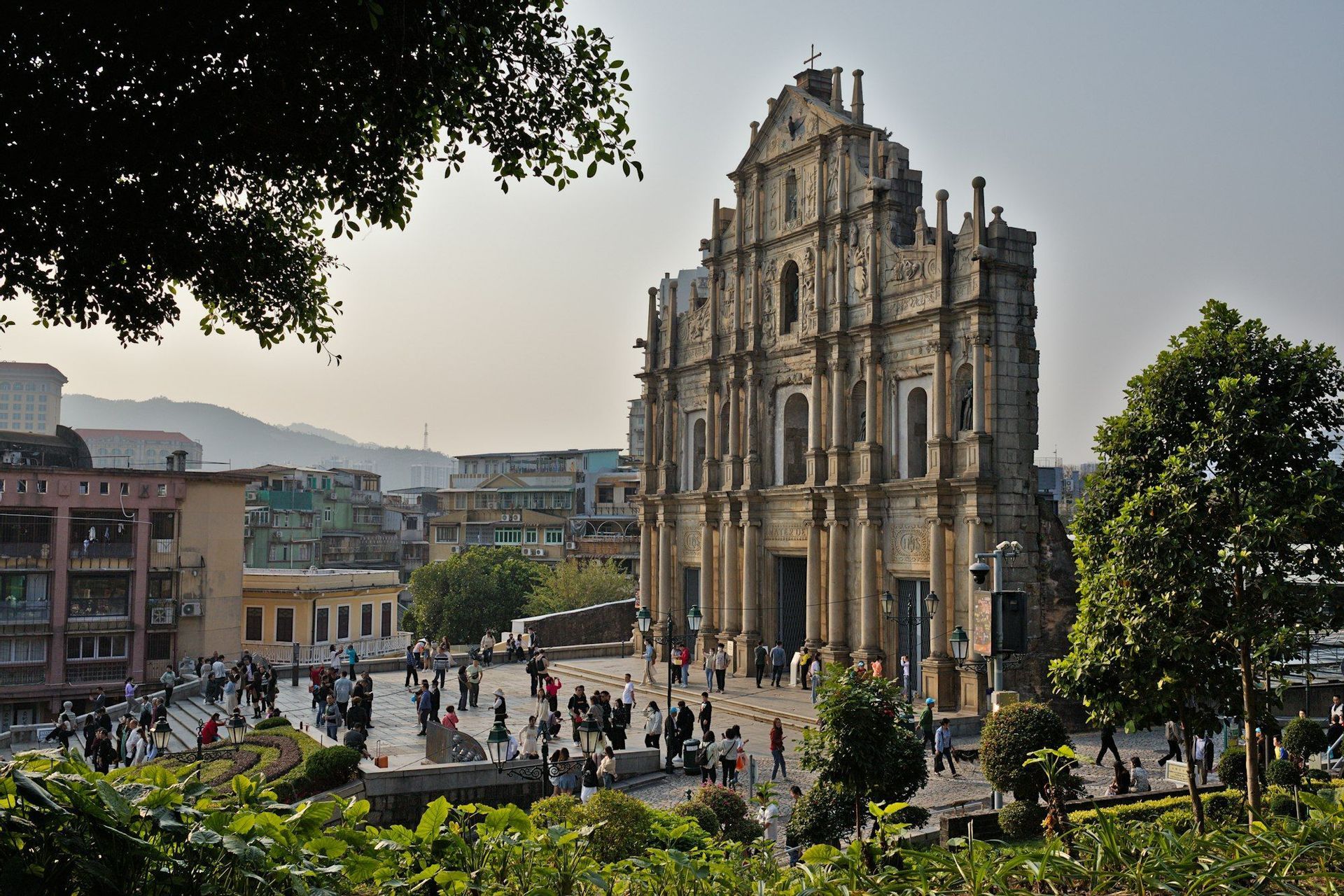 La facciata in pietra ornata di una chiesa in rovina si erge sopra una piazza affollata, con edifici cittadini e colline visibili sullo sfondo.