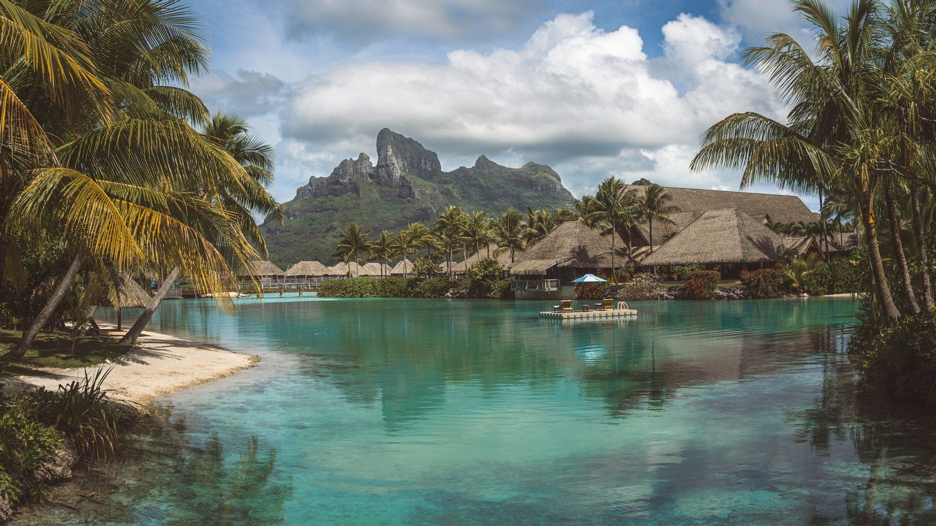 Un resort tropicale con bungalow sull'acqua e palme lungo una laguna turchese, con una montagna verde sullo sfondo.