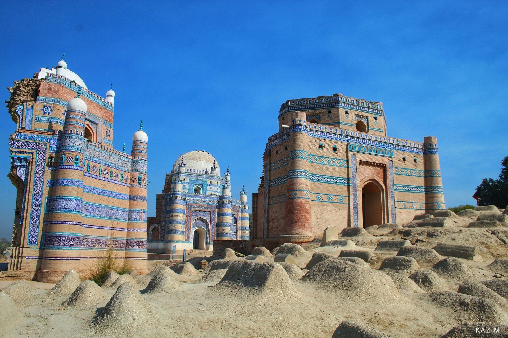 Diverse grandi tombe in mattoni, ornate con piastrelle blu, si stagliano su un campo di sepolture in terra, sotto un cielo azzurro sereno.