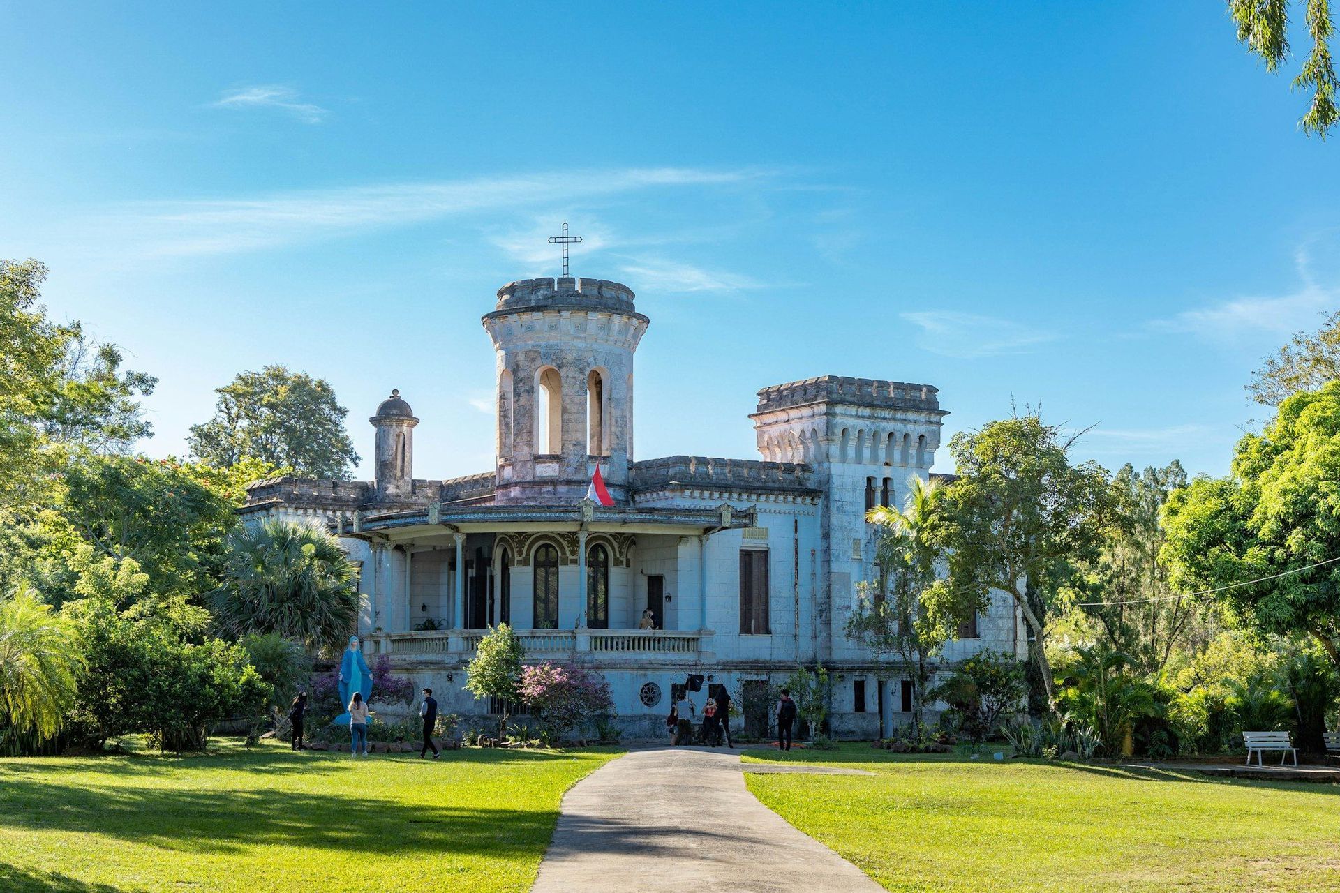 Un palazzo in pietra con più torri si erge su un grande prato verde circondato da alberi, con un sentiero che conduce all'ingresso sotto un cielo soleggiato.