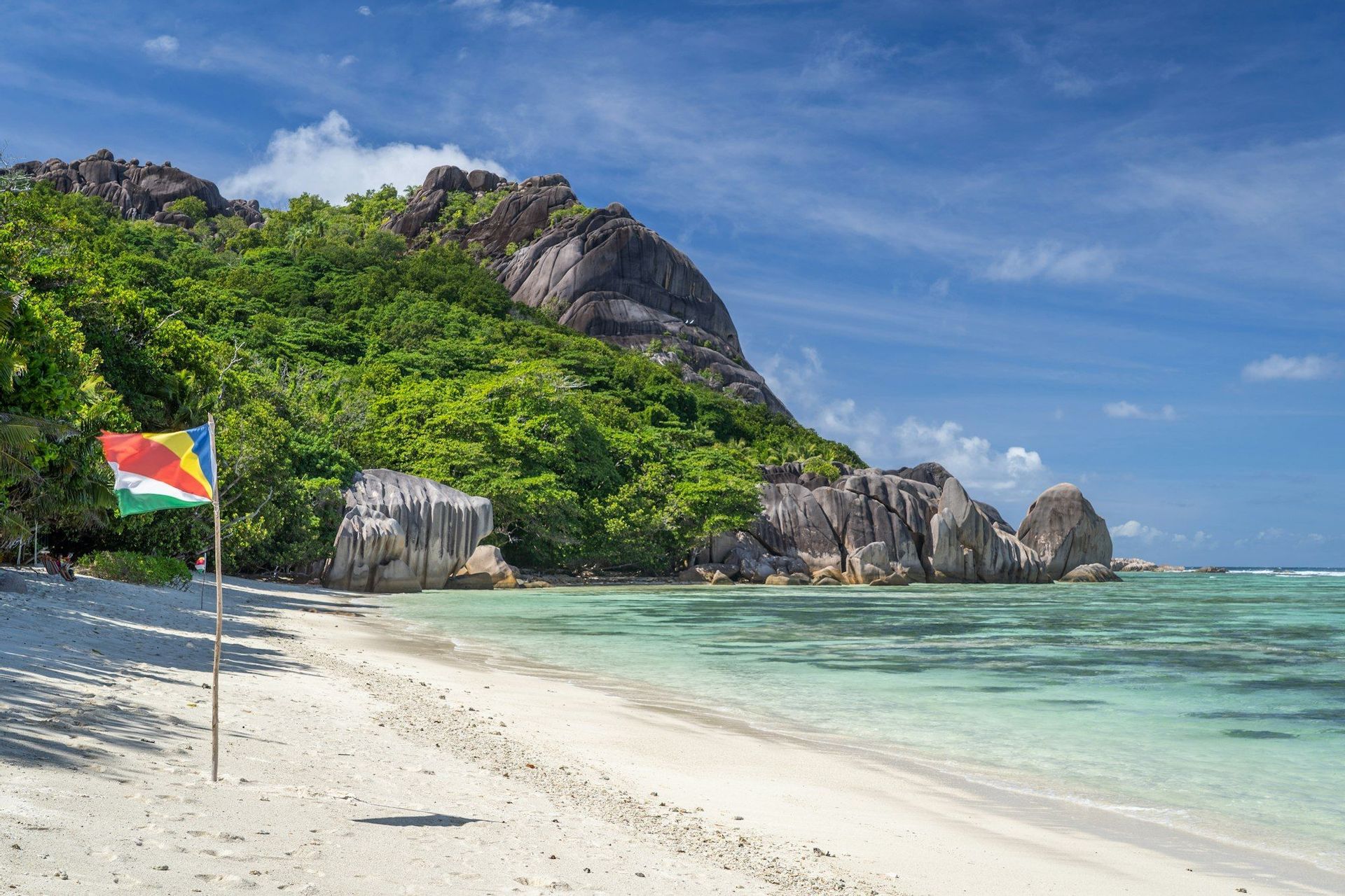 La bandiera delle Seychelles si erge su una spiaggia di sabbia bianca accanto all'acqua turchese, con una collina rocciosa e alberata sullo sfondo.