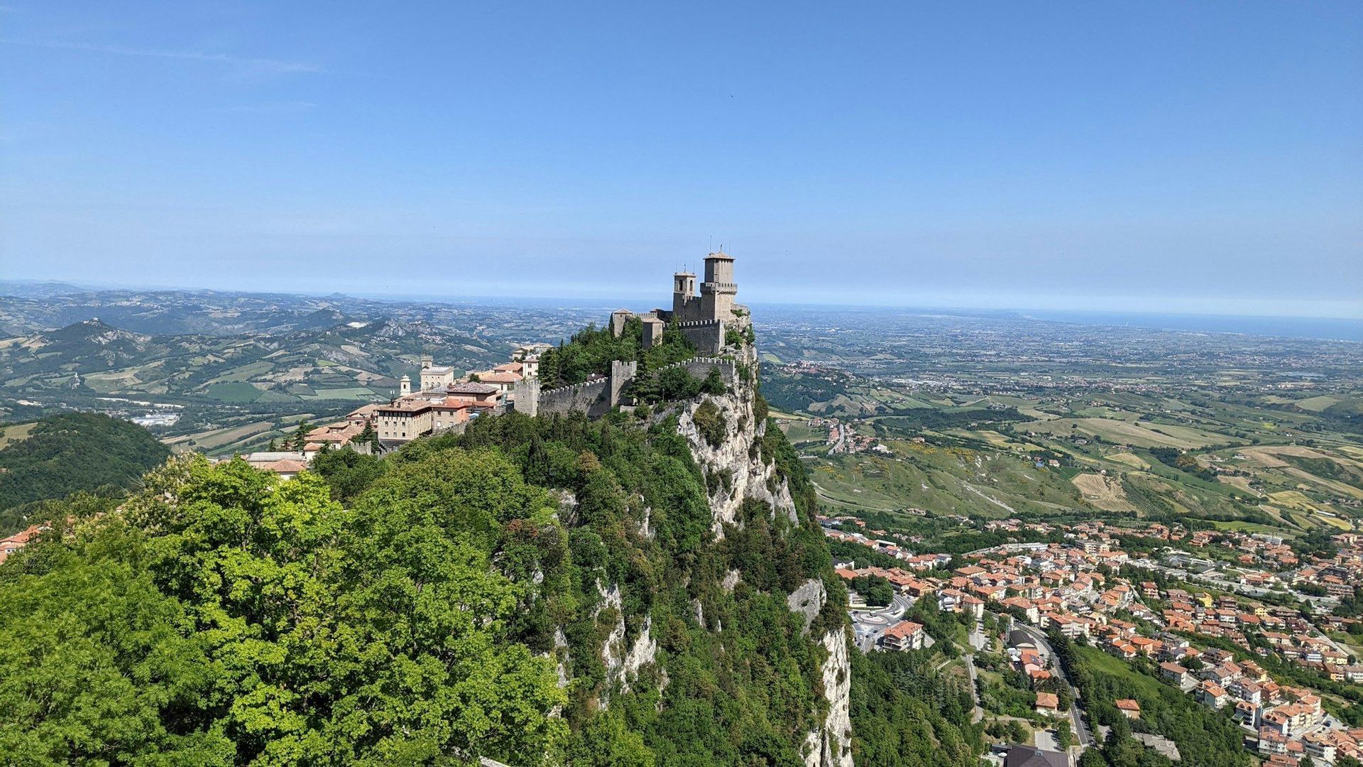 Un castello medievale con torri si erge su un'alta scogliera ricoperta di alberi, dominando un vasto paesaggio di dolci colline e città.