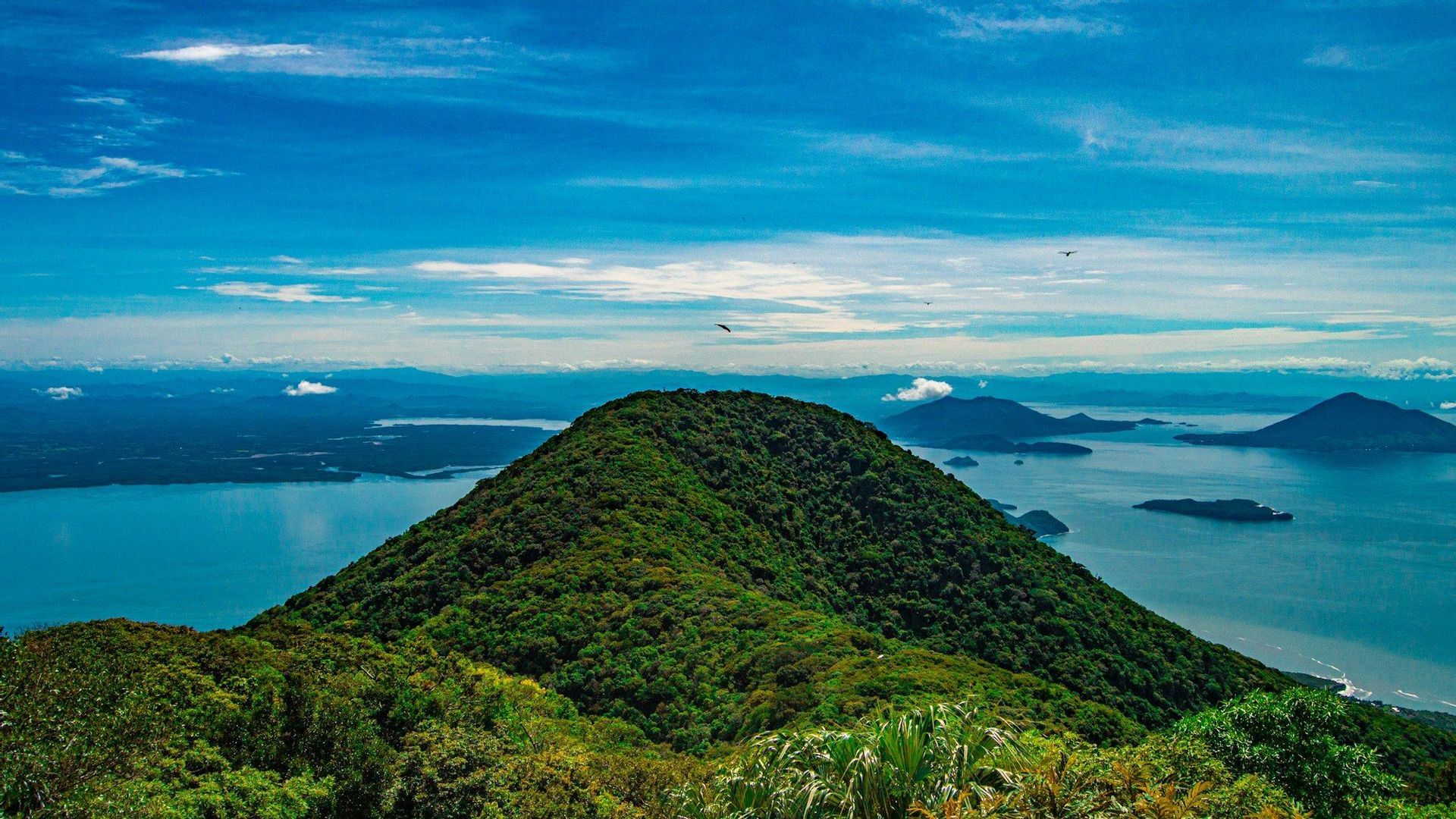 Una cima montuosa lussureggiante e verde si affaccia su un'ampia baia blu con diverse isole più piccole sotto un cielo parzialmente nuvoloso.