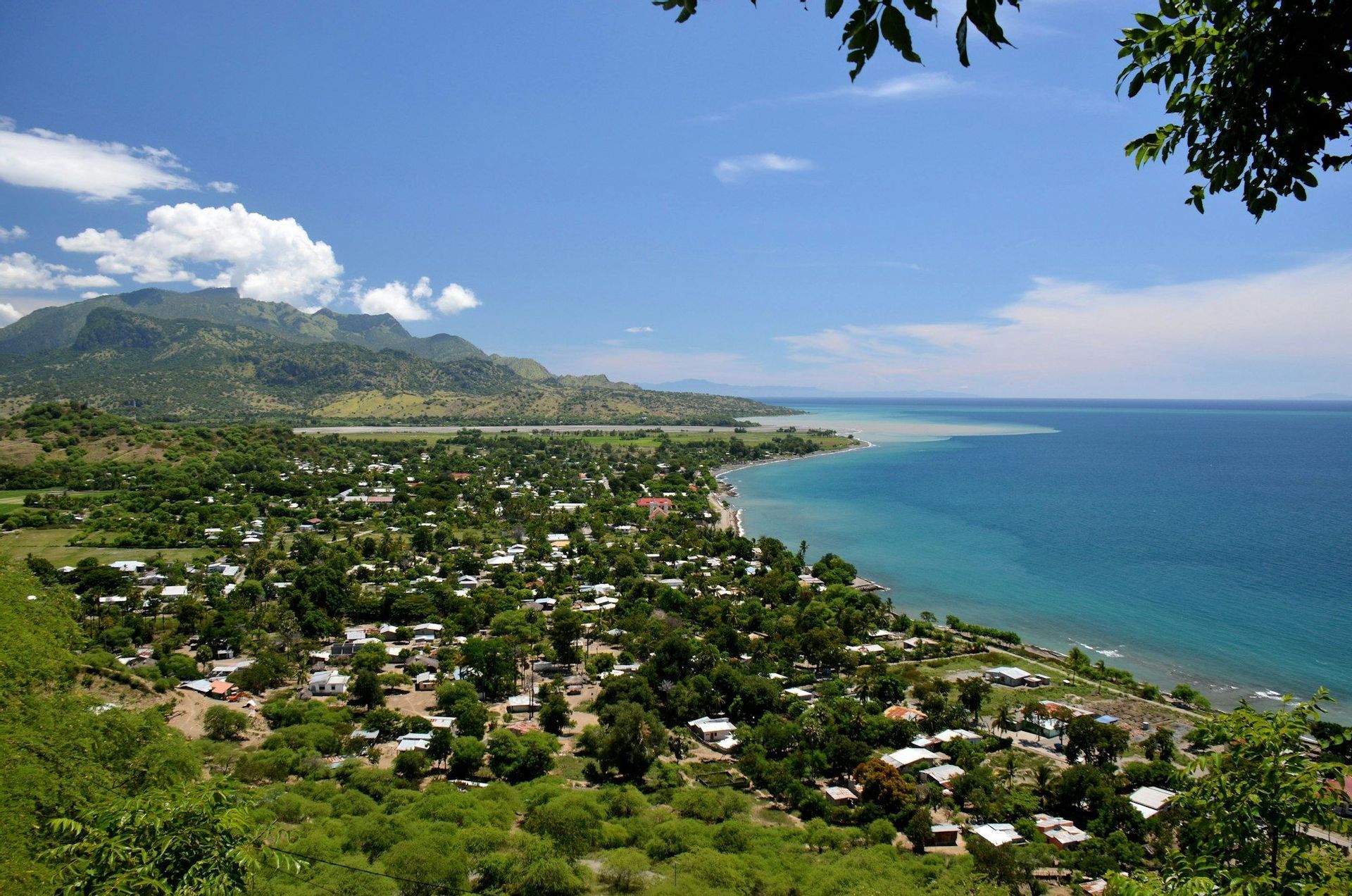 Vista dall'alto di una località costiera con vegetazione lussureggiante, incastonata tra montagne e mare azzurro sotto un cielo parzialmente nuvoloso.