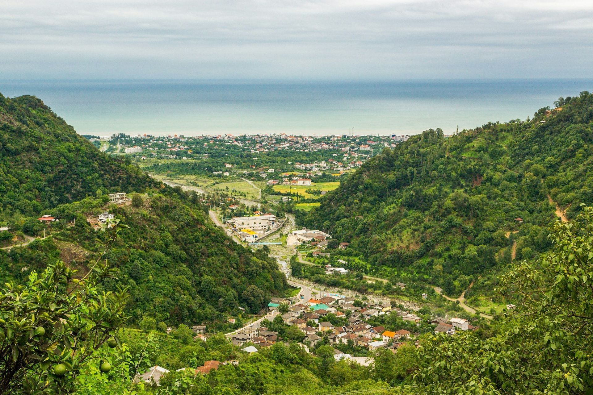 Una vista aerea su una valle lussureggiante e verde con un piccolo paese, che si estende verso una città costiera più grande e il mare.