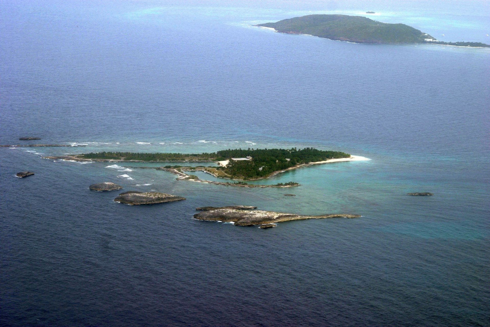 Una vista aerea di una piccola isola tropicale con edifici circondati da acqua turchese e isolotti rocciosi.