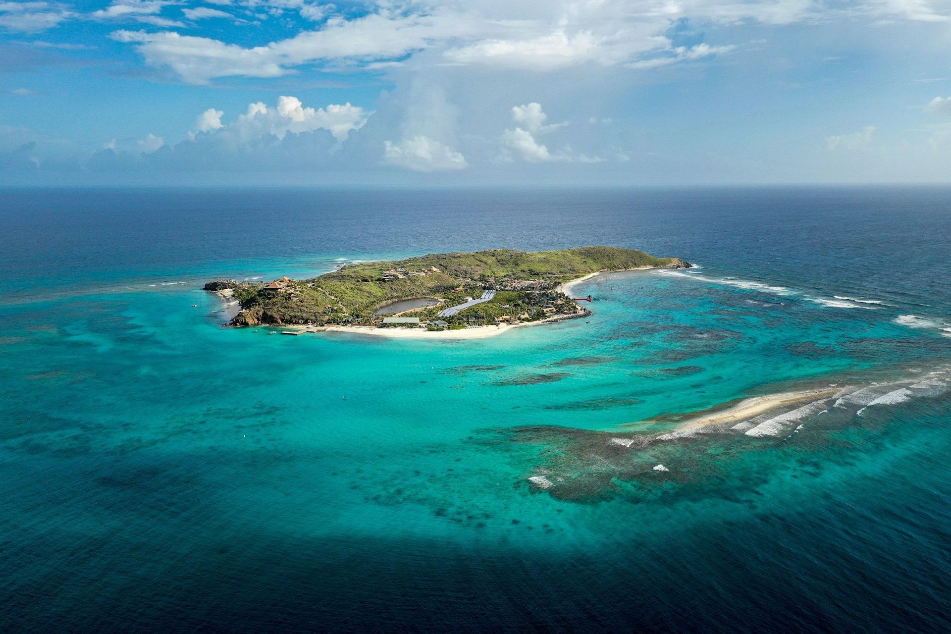 Una vista aerea di un'isola tropicale lussureggiante circondata da acque turchesi poco profonde e barriere coralline, sotto un cielo azzurro con nuvole bianche.