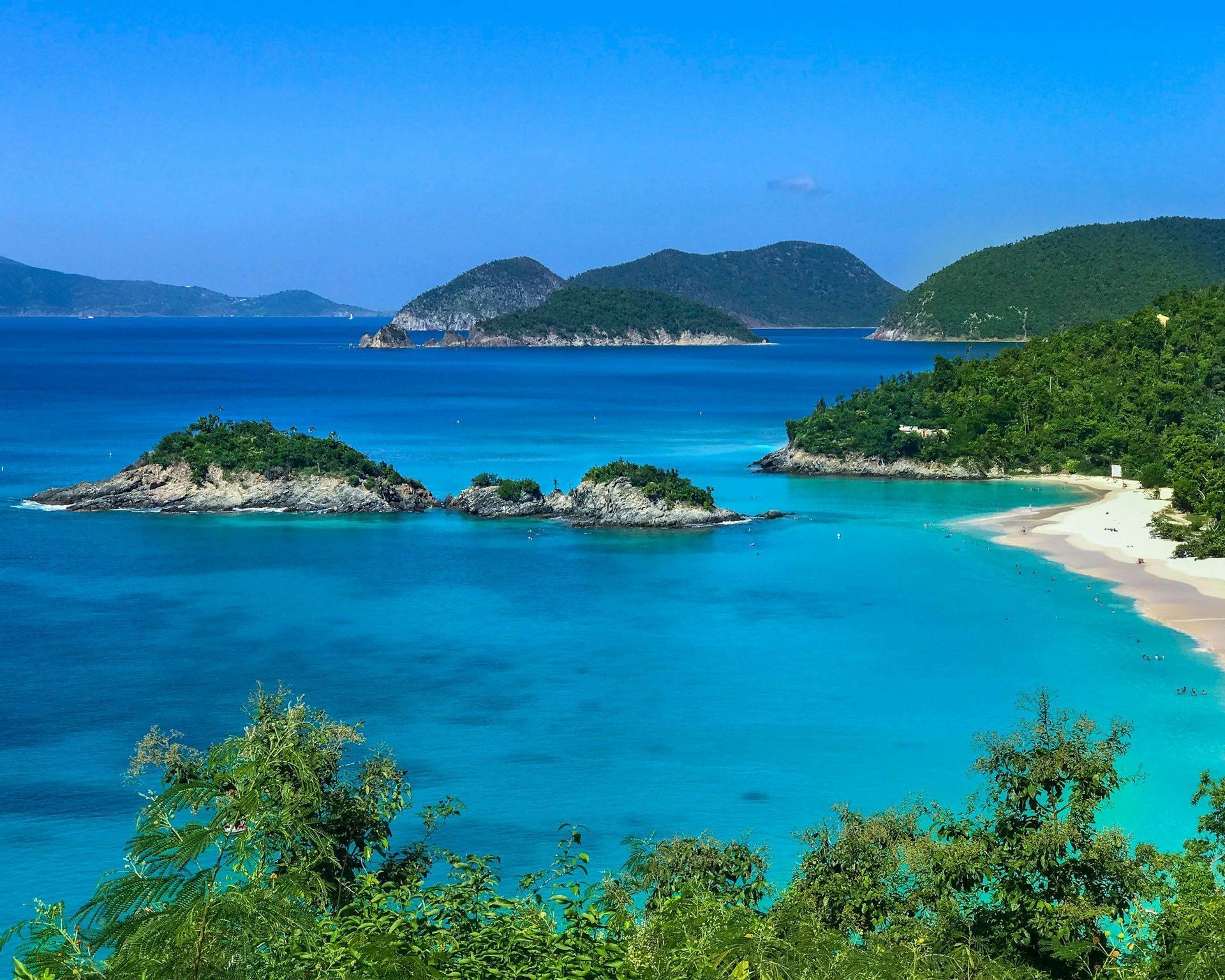 Vista dall'alto di una baia tropicale con acqua turchese, spiaggia di sabbia bianca e piccole isole verdi sotto un cielo sereno.