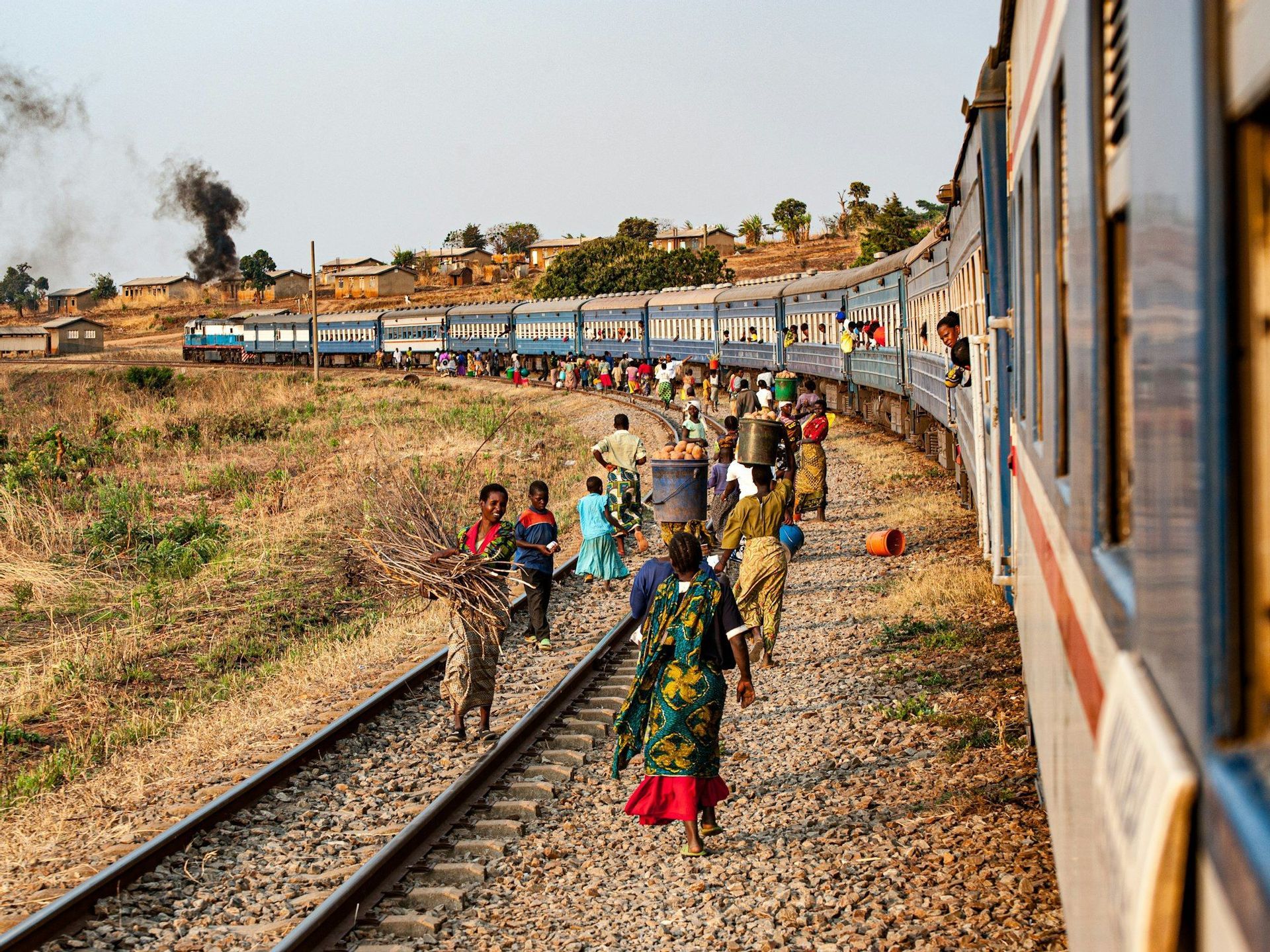 Un gruppo di persone che trasportano merci cammina sulla ghiaia accanto a un lungo treno passeggeri blu fermo su un binario curvo in una zona rurale.