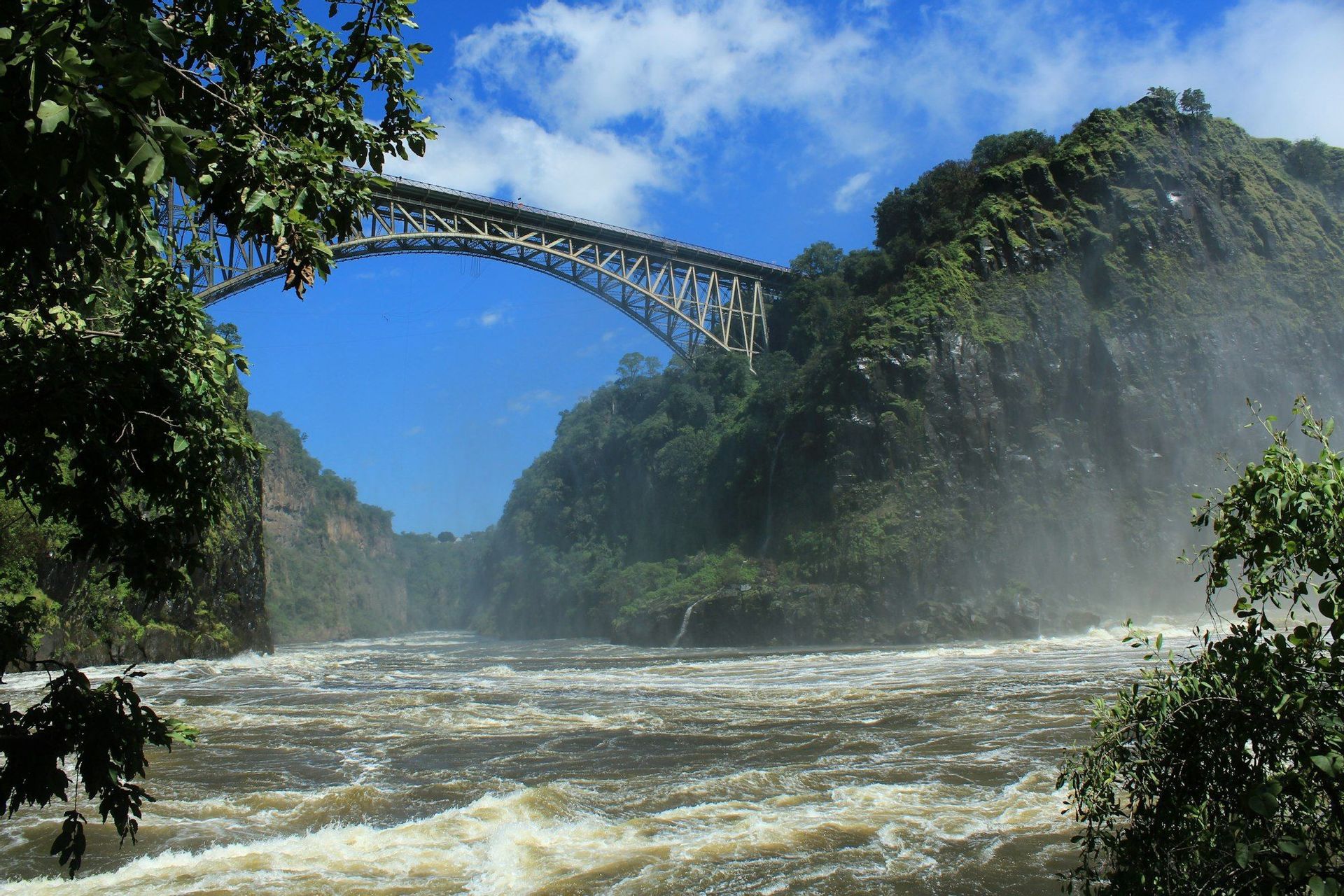 Un ponte metallico ad arco attraversa una profonda gola sopra un fiume turbolento, incorniciato da alberi verdi sotto un cielo blu.