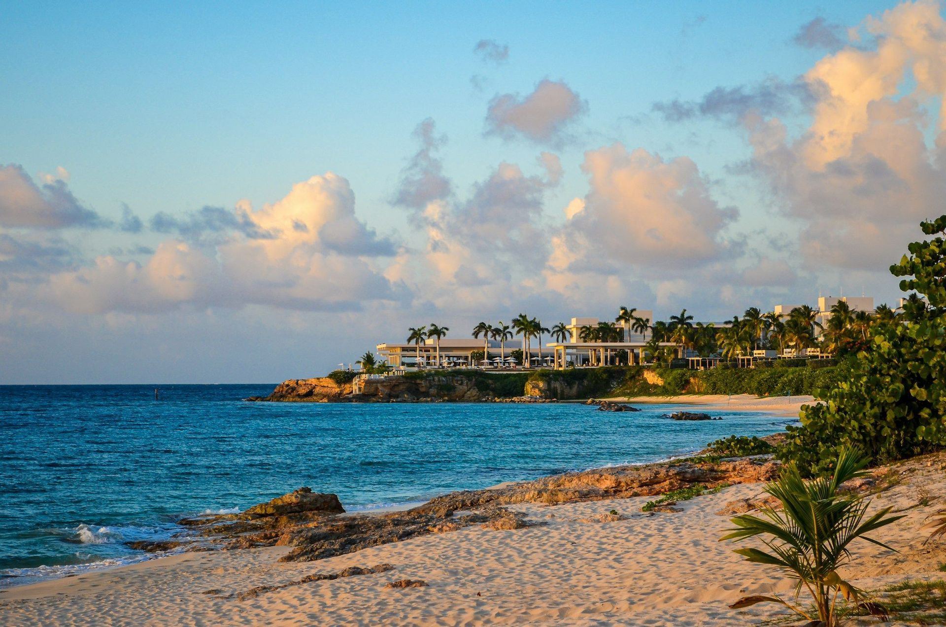 Una vista desde una playa de arena de un resort moderno con palmeras en una costa rocosa junto a agua turquesa al atardecer.