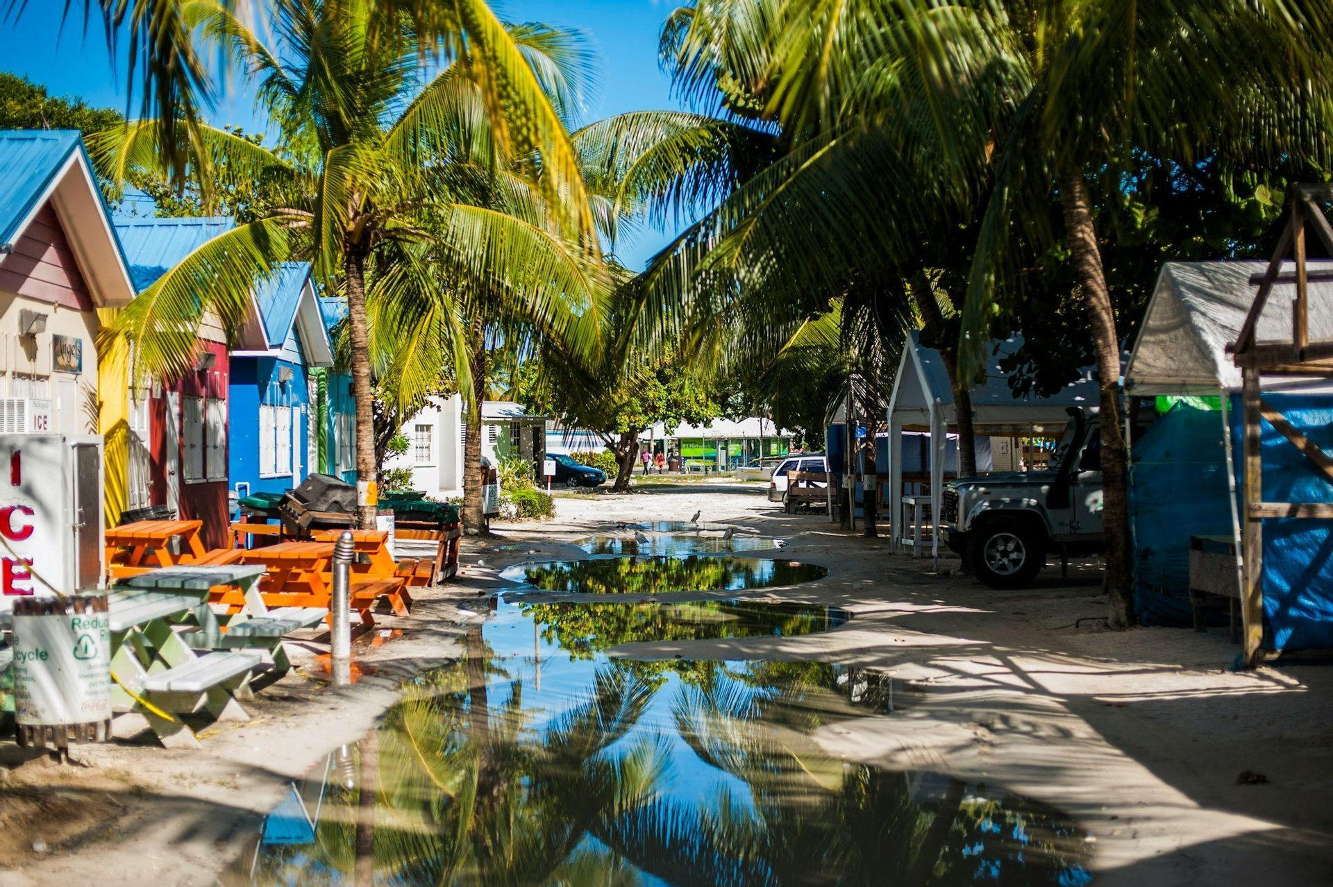 Casetas de playa coloridas y palmeras flanquean una calle de arena con grandes charcos que reflejan el cielo azul y los árboles.