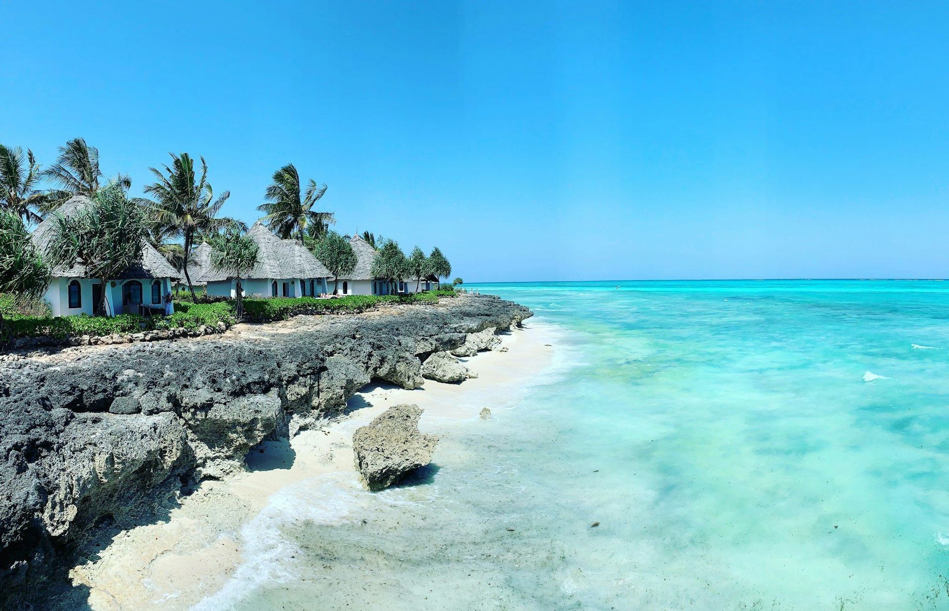 Bungalows blancos con techos de paja y palmeras bordean una costa rocosa junto a un océano turquesa bajo un cielo azul claro.