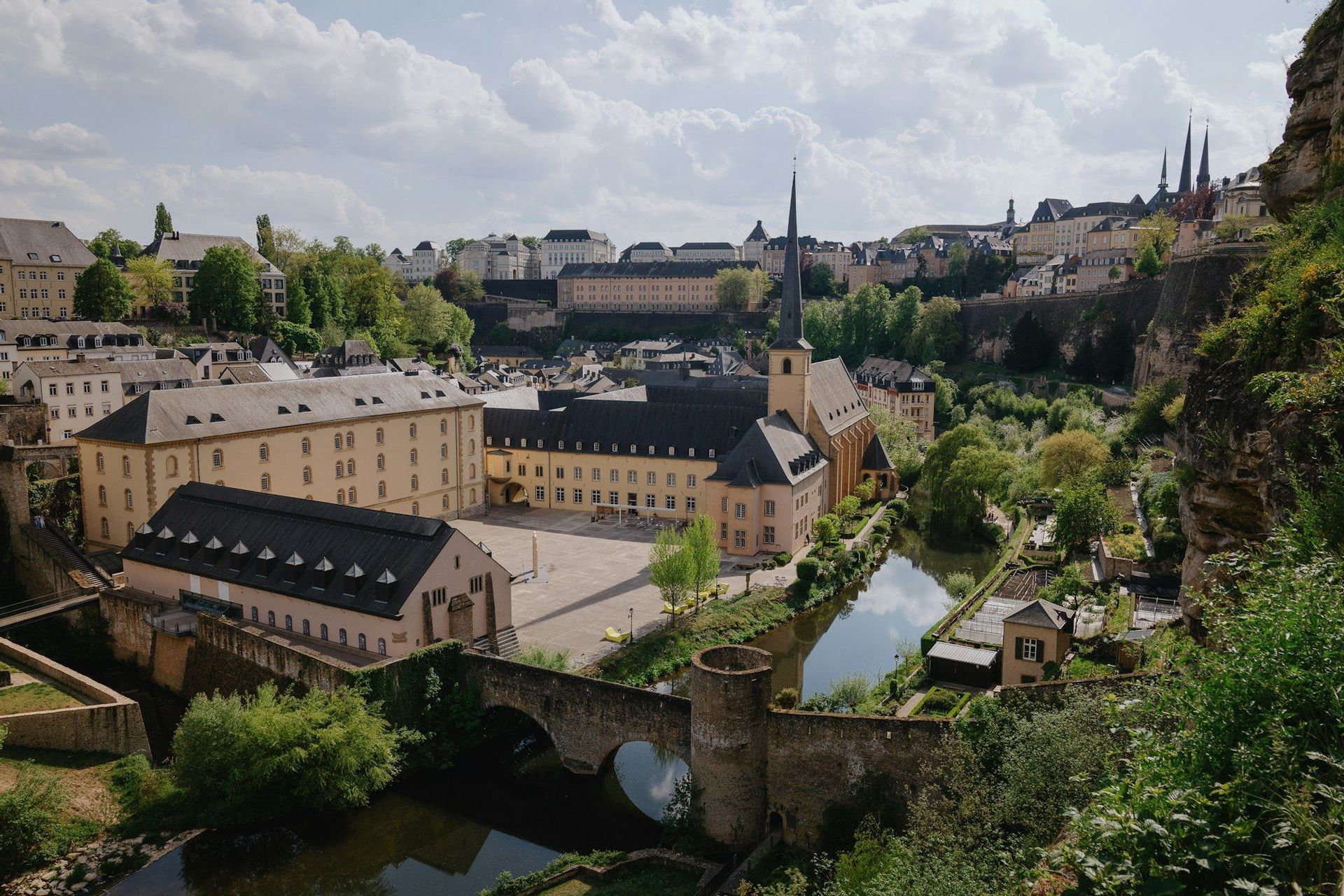 Una vista aérea de un pueblo europeo histórico con un río, un puente de piedra y edificios tradicionales bajo un cielo parcialmente nublado.