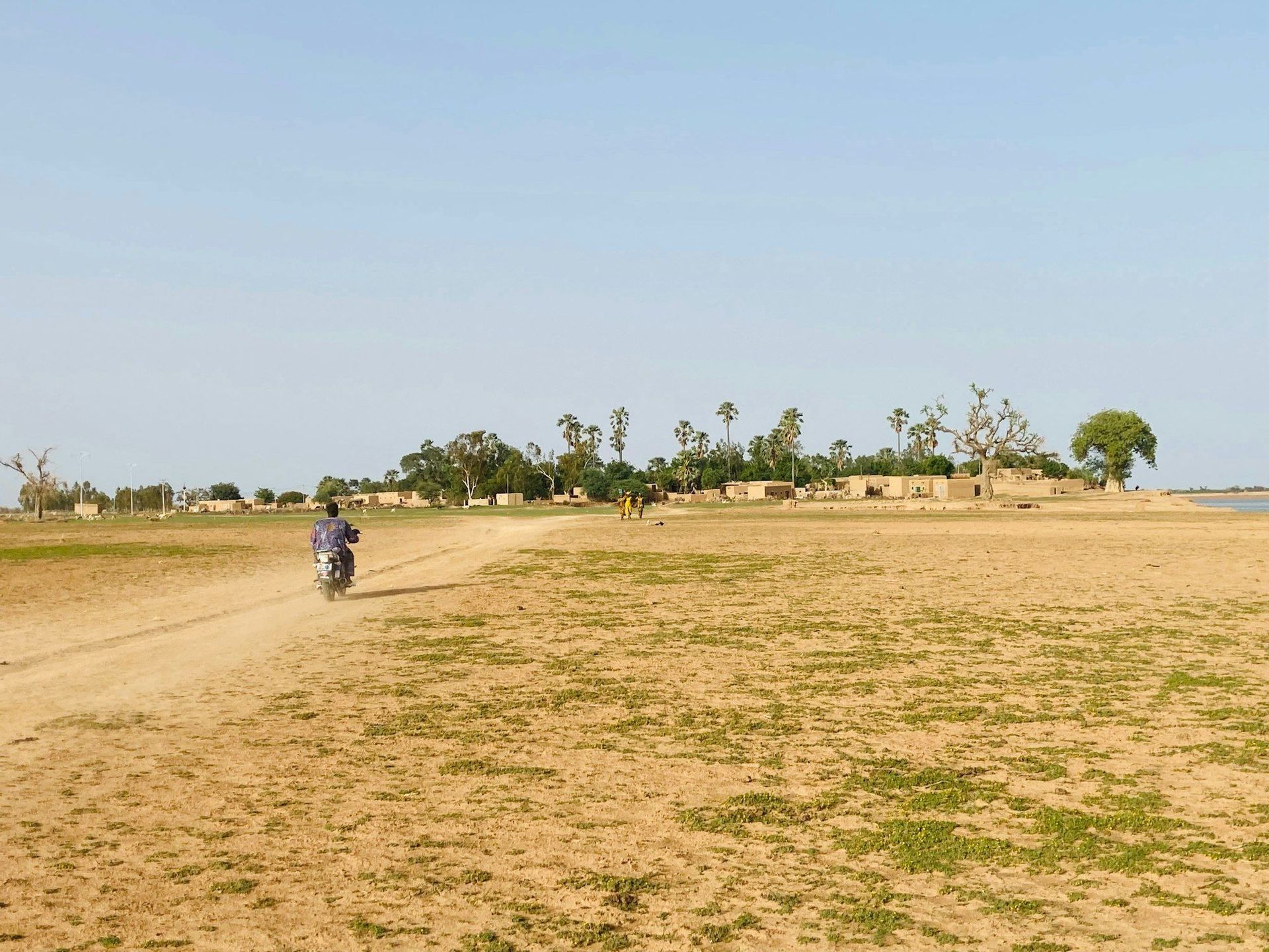 Una persona en motocicleta recorre un camino de tierra a través de un paisaje seco hacia un pequeño pueblo con palmeras.