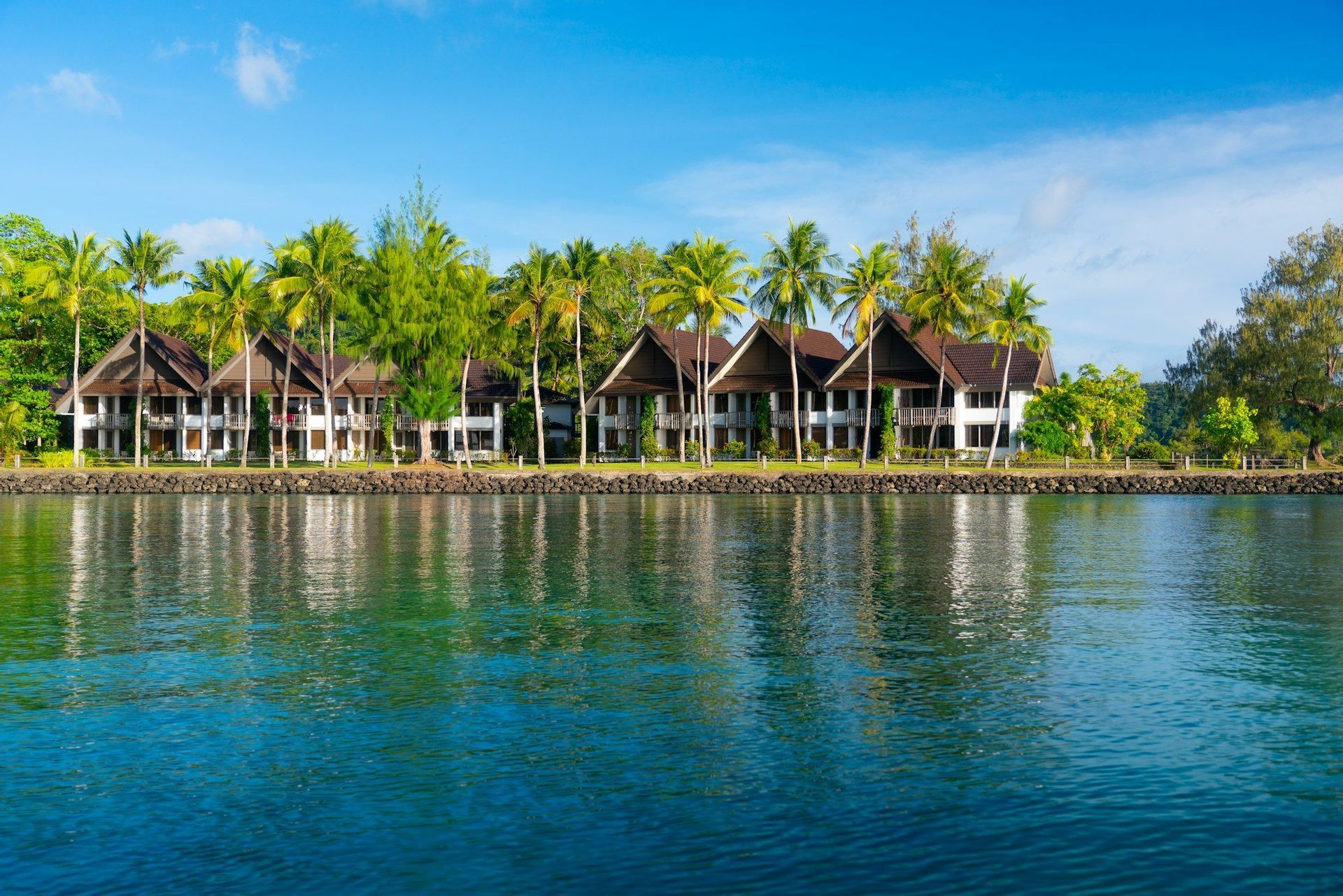 Una hilera de villas tropicales rodeadas de palmeras se refleja en el agua tranquila y azul de una laguna en un día soleado.