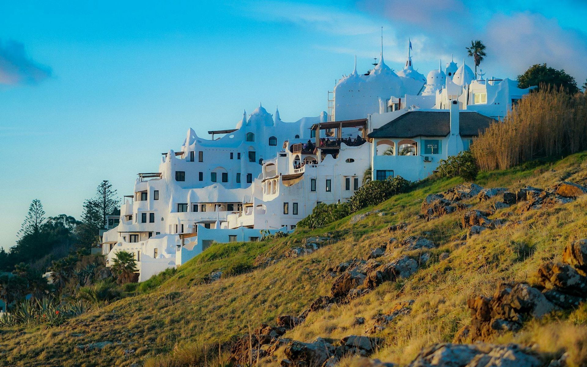 Un gran edificio blanco de varios pisos con arquitectura única se asienta en una ladera rocosa y cubierta de hierba bajo un cielo azul claro.