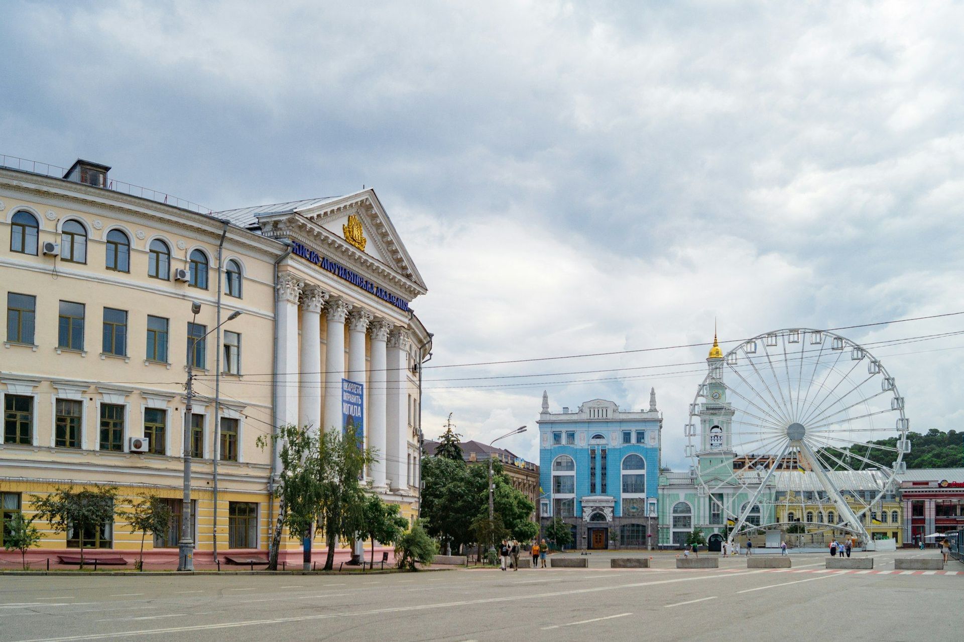 Ein weiter Stadtplatz mit einem großen Gebäude mit Säulen und einem Riesenrad vor einem bewölkten Himmel.