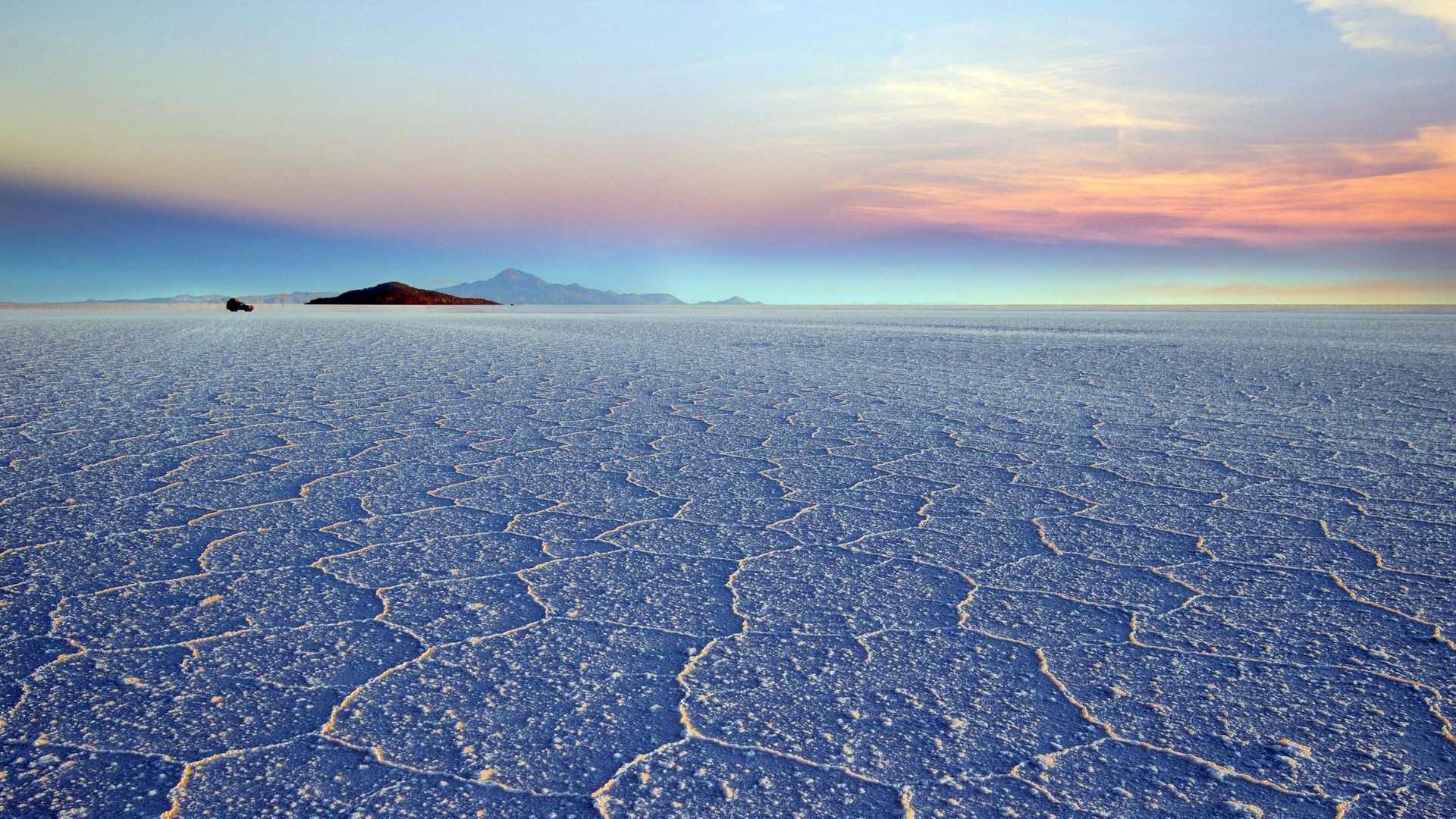 Una vasta pianura salata con motivi esagonali si estende verso montagne distanti sotto un cielo colorato all'alba.
