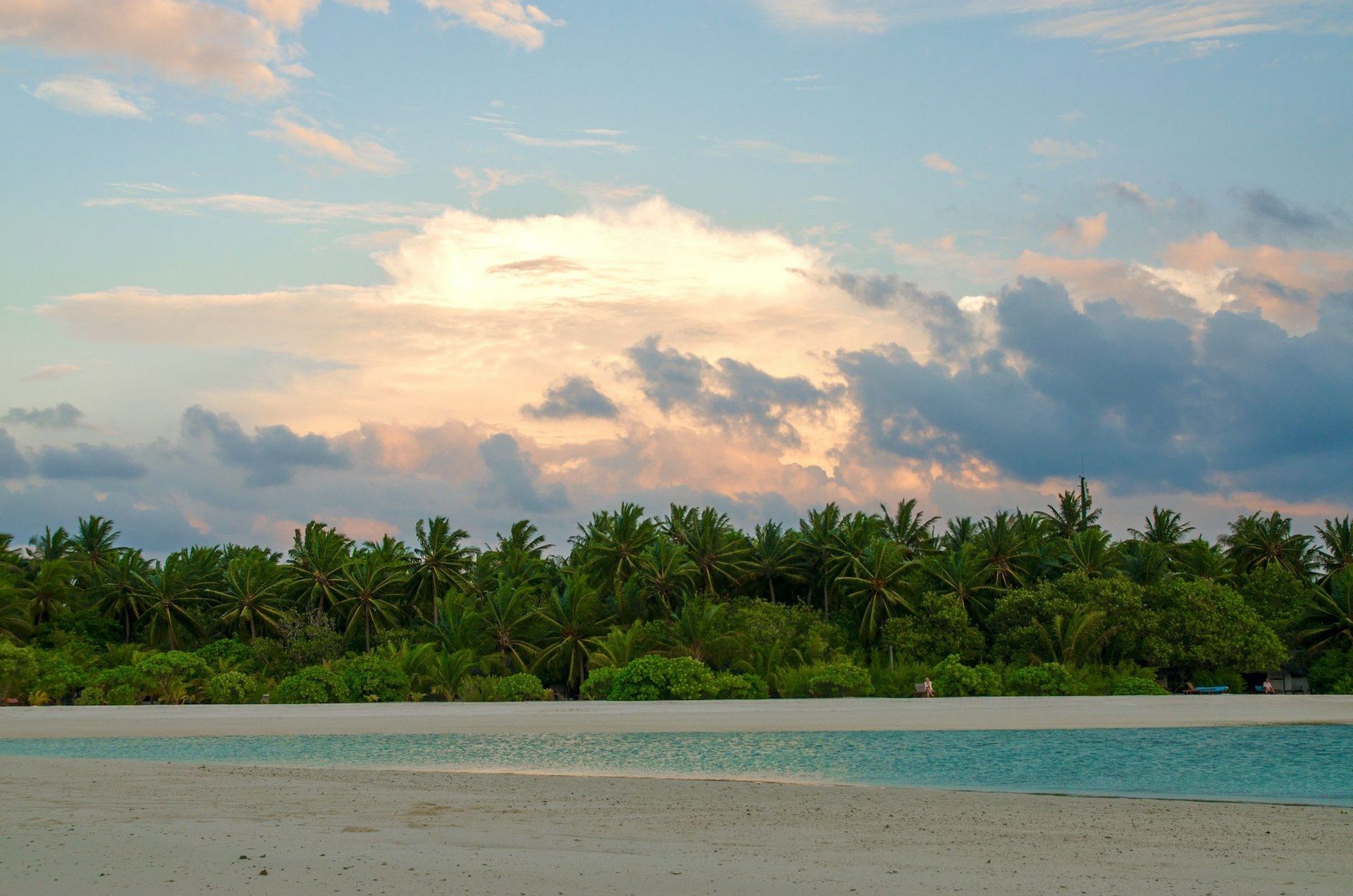 Ein Sandstrand und eine türkisfarbene Lagune vor einer dichten Palmenreihe unter einem farbenprächtigen Sonnenuntergangshimmel.
