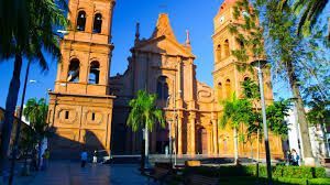 Una cattedrale di mattoni arancioni con due grandi campanili si erge dietro diverse palme sotto un cielo azzurro chiaro.