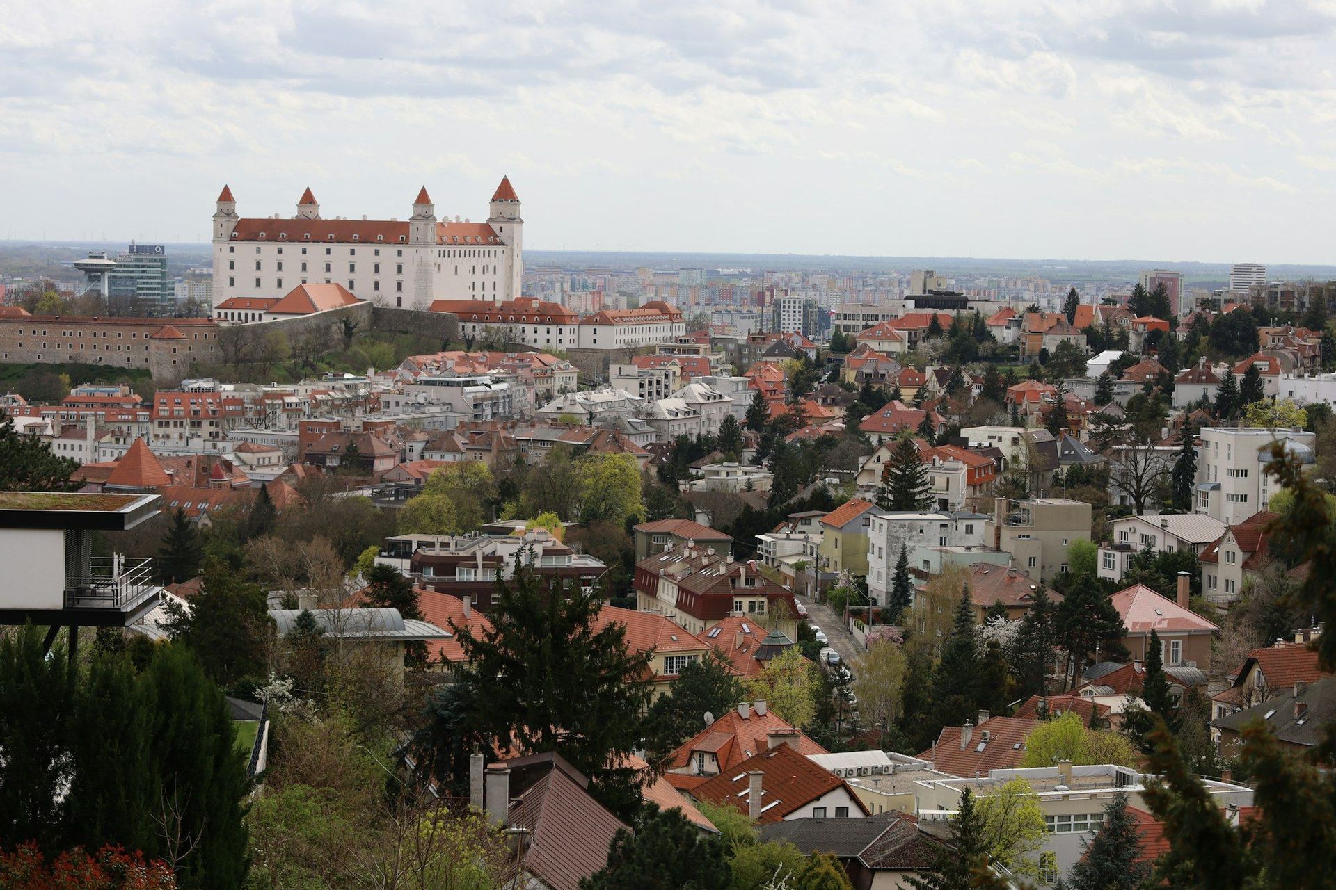Ein großes weißes Schloss mit rot gedeckten Ecktürmen thront auf einem Hügel und überblickt eine weitläufige Stadtlandschaft mit vielen Bäumen unter einem bewölkten Himmel.