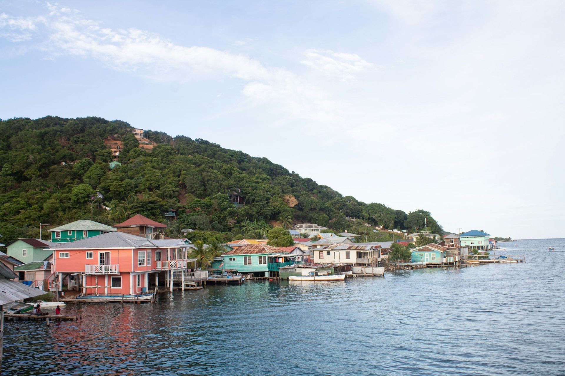 Des maisons colorées sur pilotis surplombent l'eau dans un village côtier adossé à une colline luxuriante et arborée.