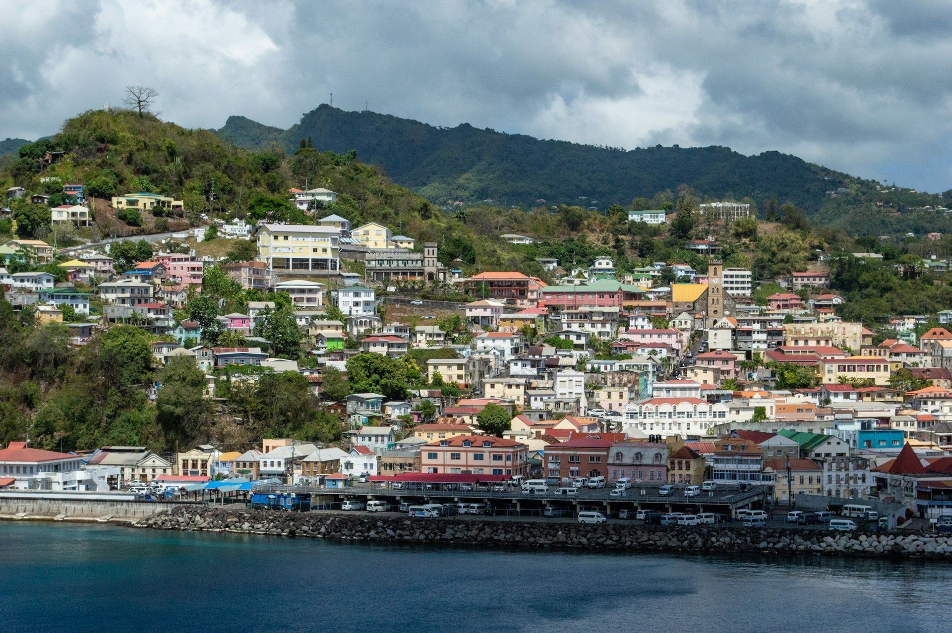 Une vue panoramique d'une ville côtière colorée construite sur une colline verte et luxuriante, avec la mer au premier plan et des montagnes à l'arrière-plan.