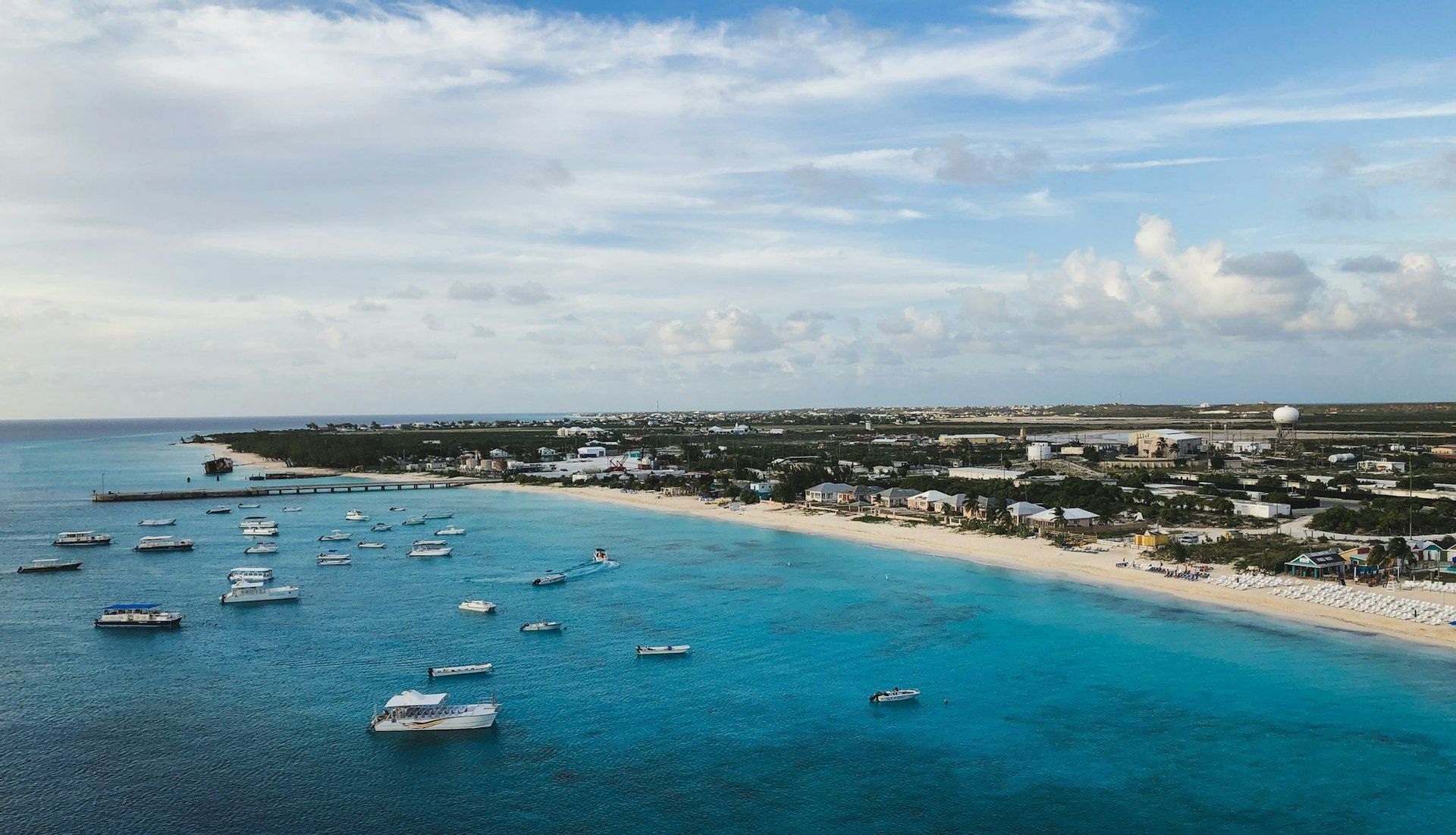 Vue aérienne de nombreux bateaux blancs ancrés dans des eaux turquoise le long d'un littoral sablonneux avec une ville en arrière-plan.