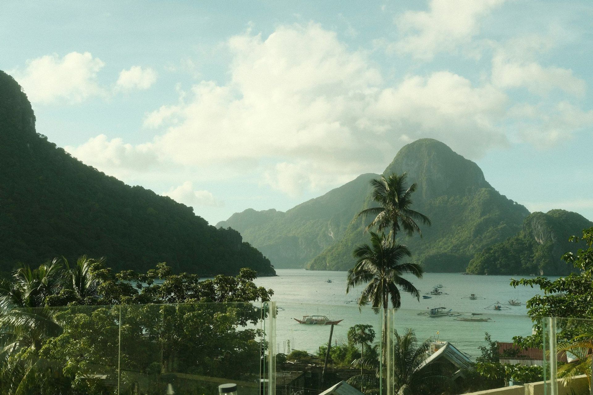 Une vue panoramique sur une baie avec des bateaux, encadrée par des palmiers et de grandes montagnes verdoyantes, sous un ciel partiellement nuageux.